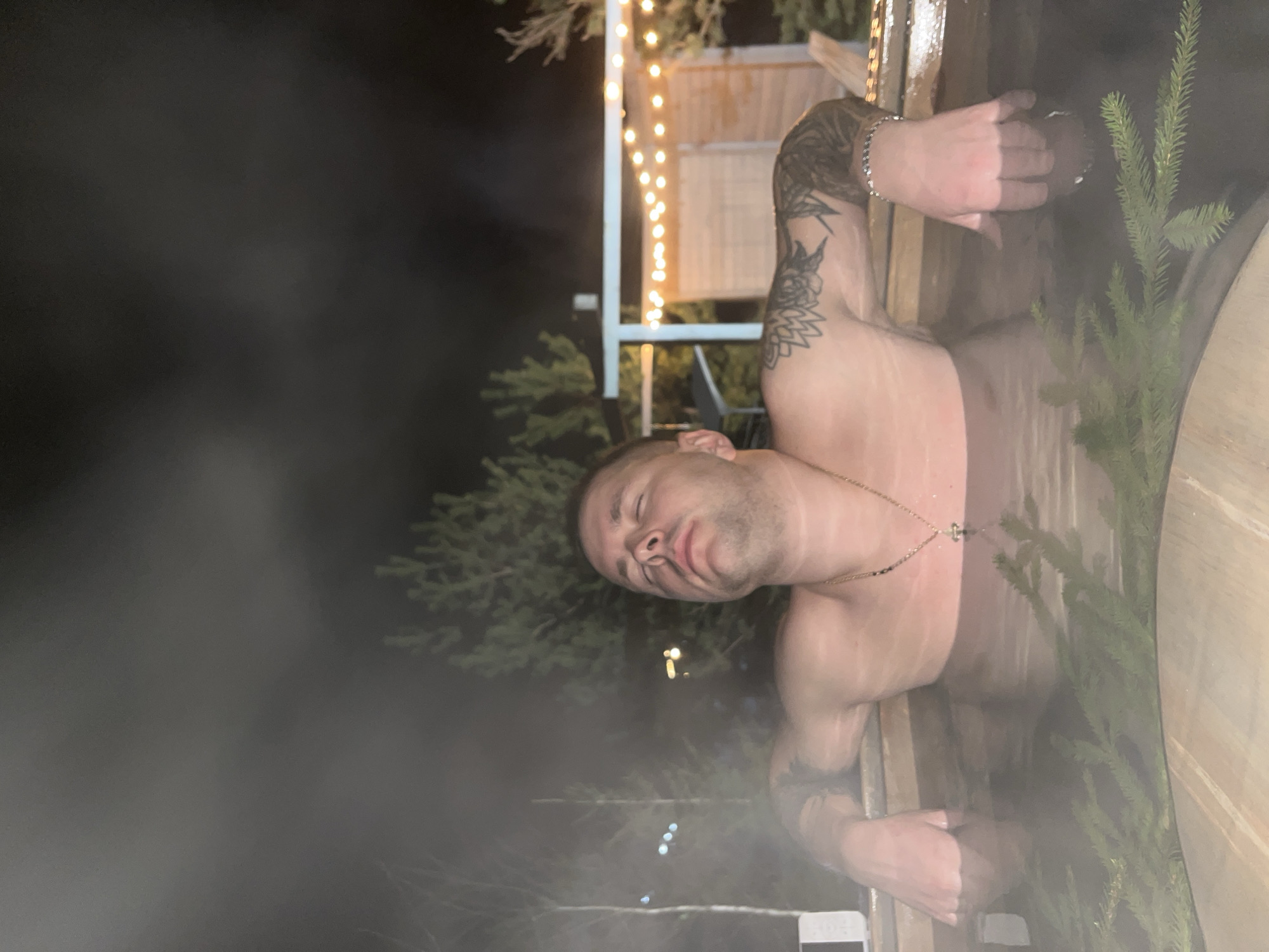 A person with tattoos is visible from the chest up in a hot tub or barrel-style sauna. The person has short hair and is wearing a gold chain around their neck. Vapor rises from the water. Green conifer branches are placed inside the hot tub. Behind the person is a building with string lights hanging from it, suggesting it is nighttime. The location is likely Литвиново, Russia.