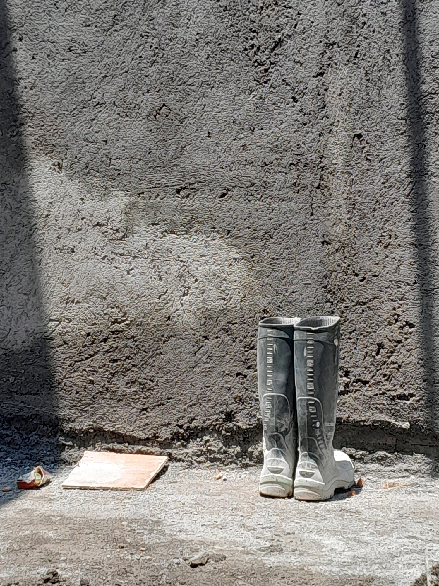 A pair of dark grey, knee-high rubber boots with white soles and accents are positioned side-by-side on a concrete ground in front of a rough, textured grey concrete wall. The boots appear worn, with some scuff marks visible. A single light brown tile, possibly a sample or discarded building material, lies on the ground to the left of the boots, partially in shadow. To the far left, a crumpled piece of red and silver material, possibly a wrapper, is also visible. The wall is unevenly textured and shows patches of lighter and darker grey, indicating it is likely unfinished or weathered. Sunlight casts a diagonal shadow across the upper portion of the wall from the left. The ground is a mix of concrete and dirt, with some small debris scattered around. The overall scene suggests a construction or work site. The location is identified as Pakisaji, Indonesia.