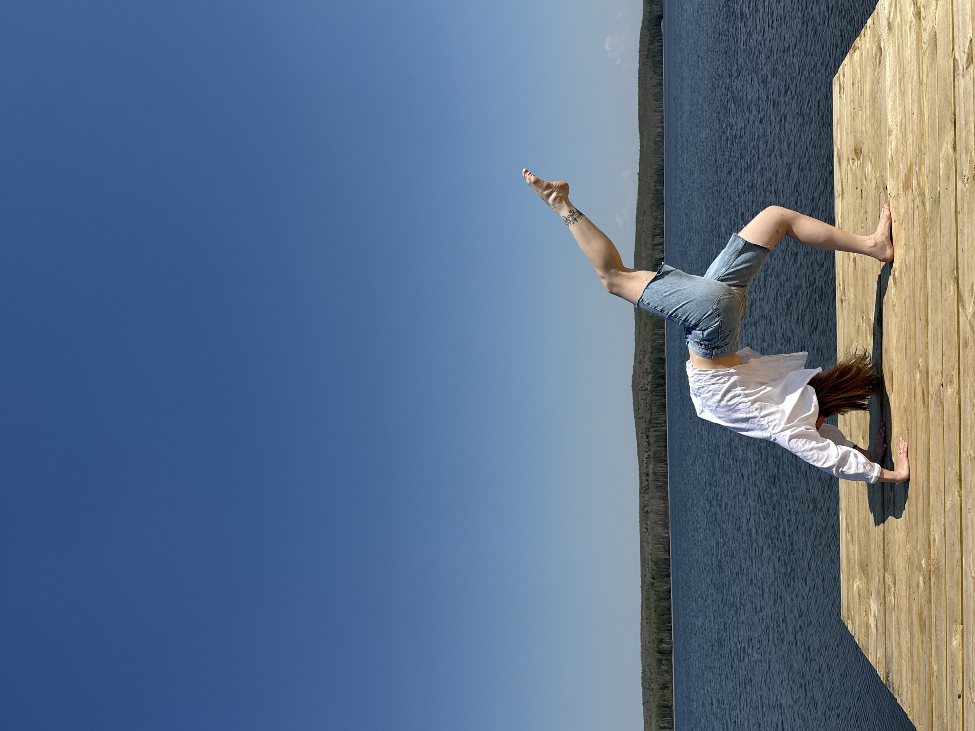 A woman in a white long-sleeved shirt and denim shorts is in a yoga backbend on a wooden dock near a lake and wooded shoreline under a blue sky. The photograph is rotated clockwise by 90 degrees. The location is likely near a body of water in Chaikovsky, Russia.