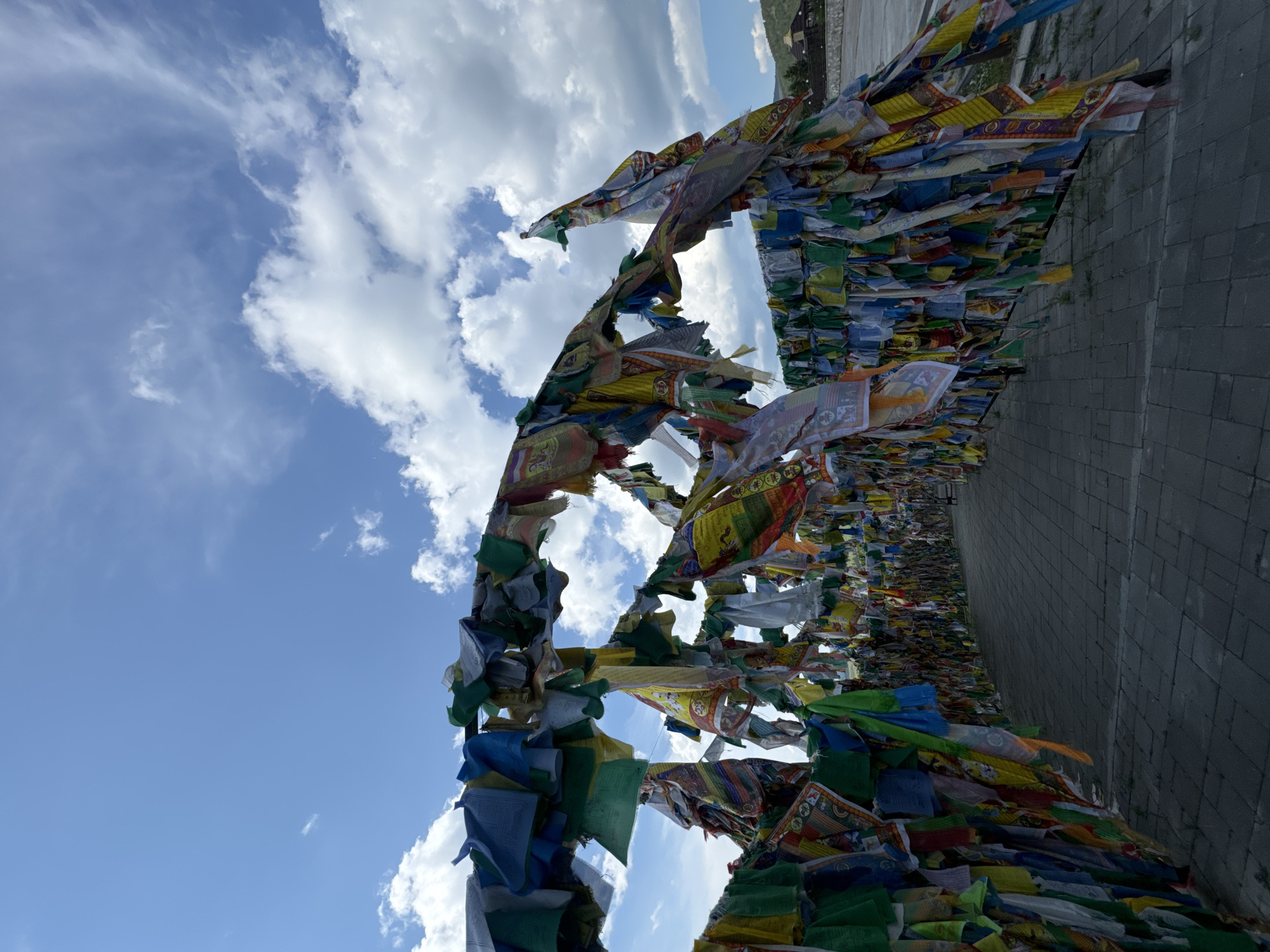 The image shows a pathway lined with colorful prayer flags. The flags are strung across the pathway, creating an arch-like effect. The pathway is paved with stone tiles. In the background, there are some buildings and a distant hill. The sky is blue with white clouds. This is the Ivolginsky Datsan, a Buddhist monastery located in the Republic of Buryatia, Russia, near Ulan-Ude.
