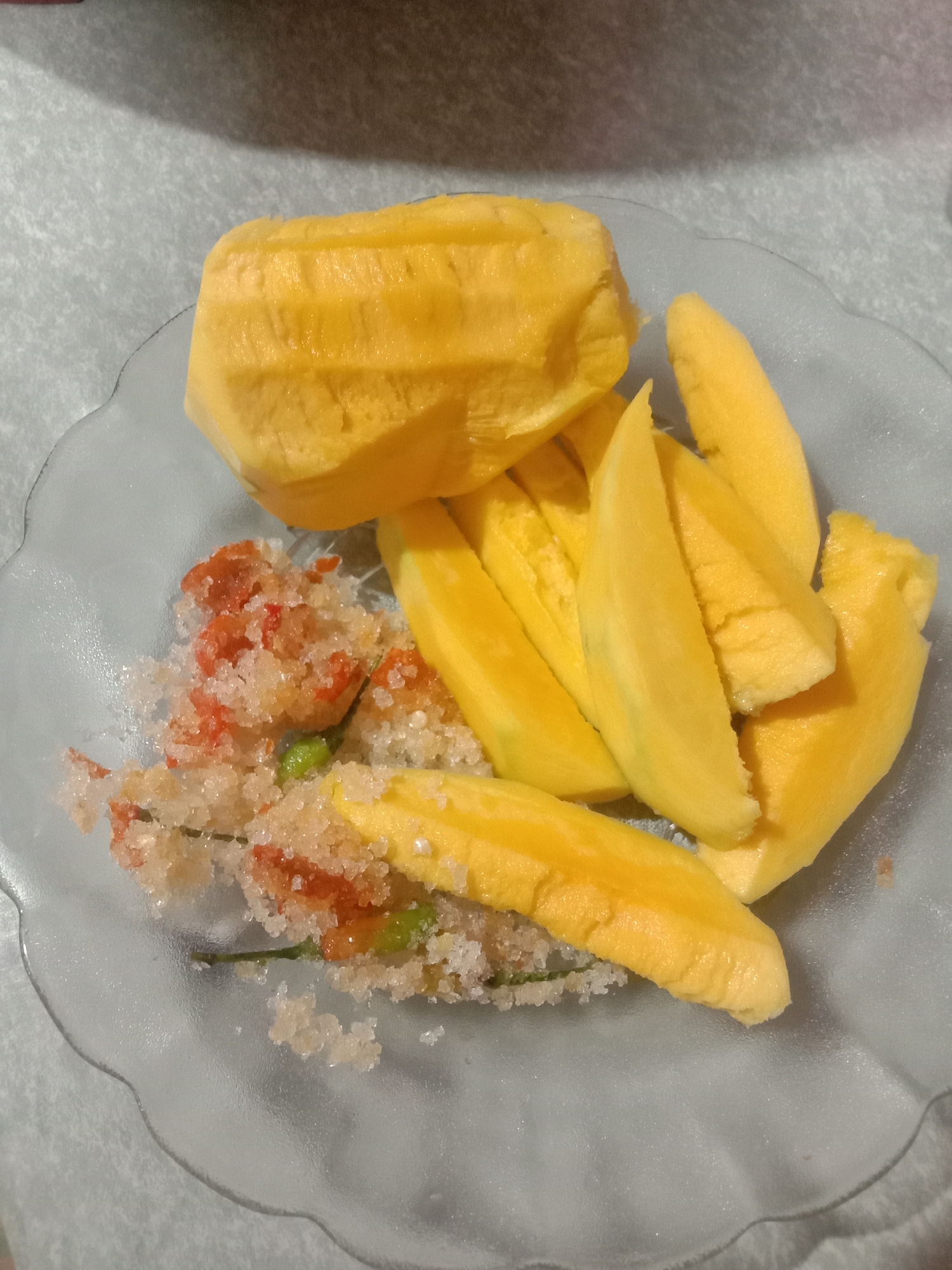 A plate of peeled mangoes is on a table. The mangoes are sliced, except for one large section that is intact. The mangoes are accompanied by a mixture of salt, red pepper, and green chili. The plate is clear glass with a scalloped edge. The table has a light-colored, textured surface. The location is Lhokjok, Indonesia. No people are visible.