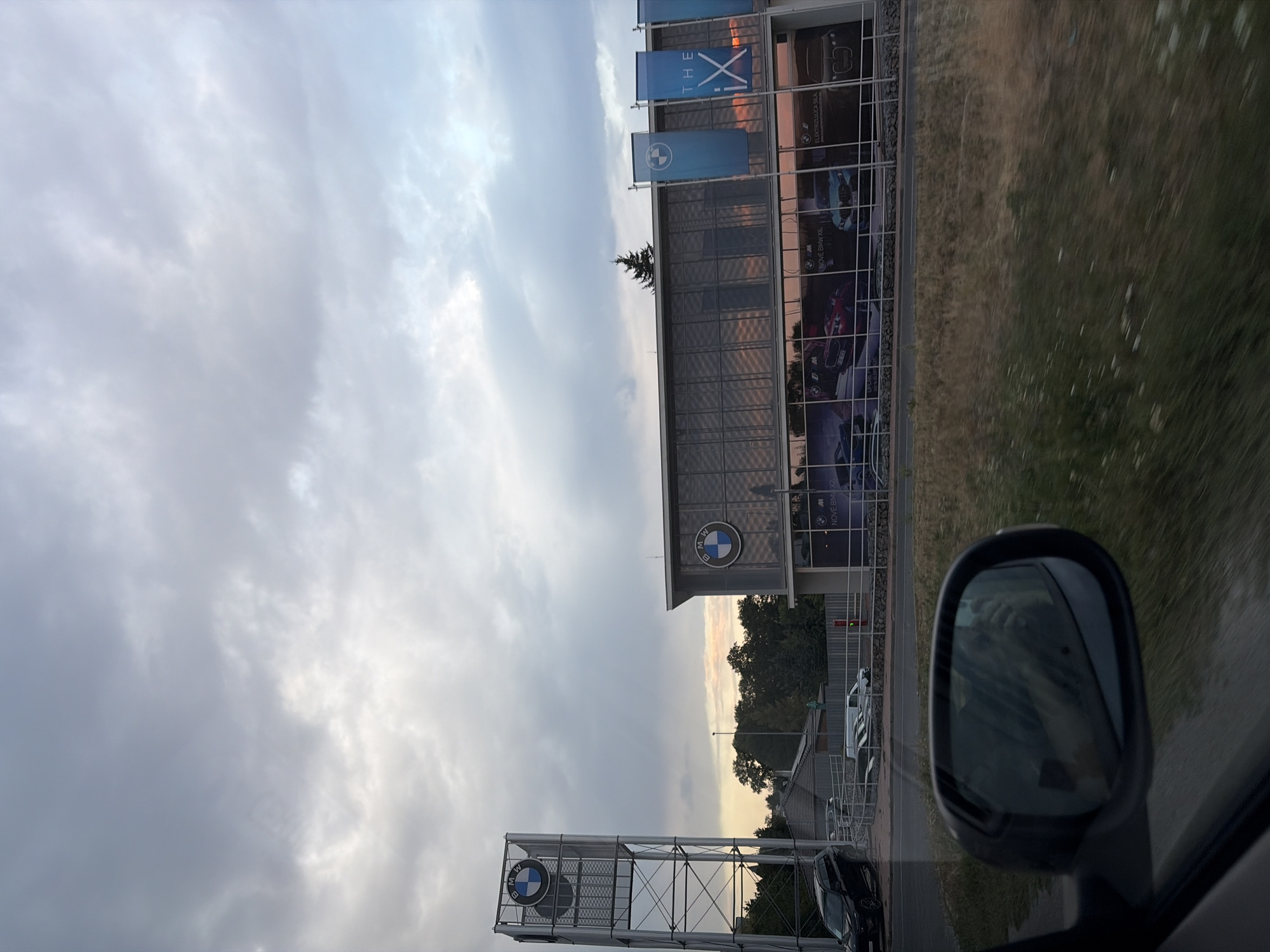 The image captures a BMW dealership in Nitra, Slovakia, under a cloudy sky. The dealership's modern architecture features a glass facade reflecting the sky. A tall metal tower with a prominent BMW logo stands to the left, while banners with "THE iX" are displayed on the building's exterior. Several cars are visible in front of the dealership, and a dark SUV is parked to the left. The view is from the perspective of a vehicle, with a side mirror reflecting a person's hand holding a phone. The foreground shows a grassy area with some dry vegetation and small white flowers.