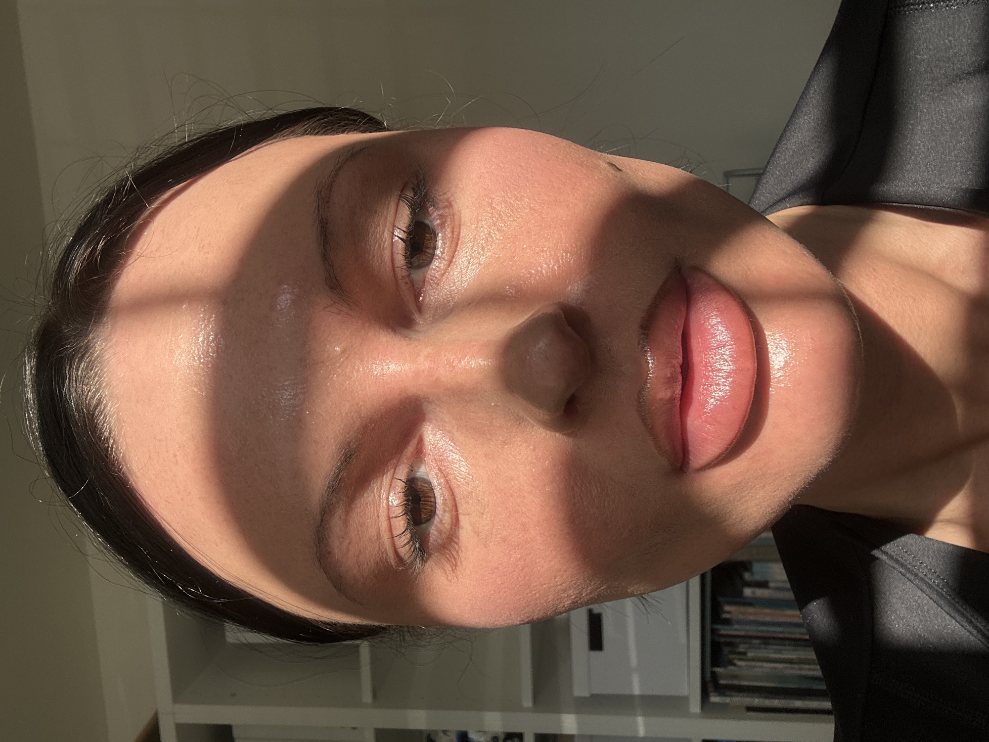 A close-up, front-facing photographic portrait of an individual in an indoor setting within Wealden, United Kingdom. The subject, identified as likely female, occupies the majority of the frame, from the forehead to the upper chest. Direct, strong sunlight from the viewer's right illuminates the face, casting pronounced, elongated shadows across the forehead, both eyes, the nose bridge, and the chin. The subject has dark hair pulled back, dark brown irises, and full, slightly parted lips exhibiting a pink hue and a reflective sheen. The skin across the face also shows a noticeable sheen. A dark-colored garment is visible around the neck and shoulders. The background on the left is blurred, revealing a white shelving unit containing white storage boxes and a collection of books or binders. The background on the right is an indistinct, plain light-colored wall.