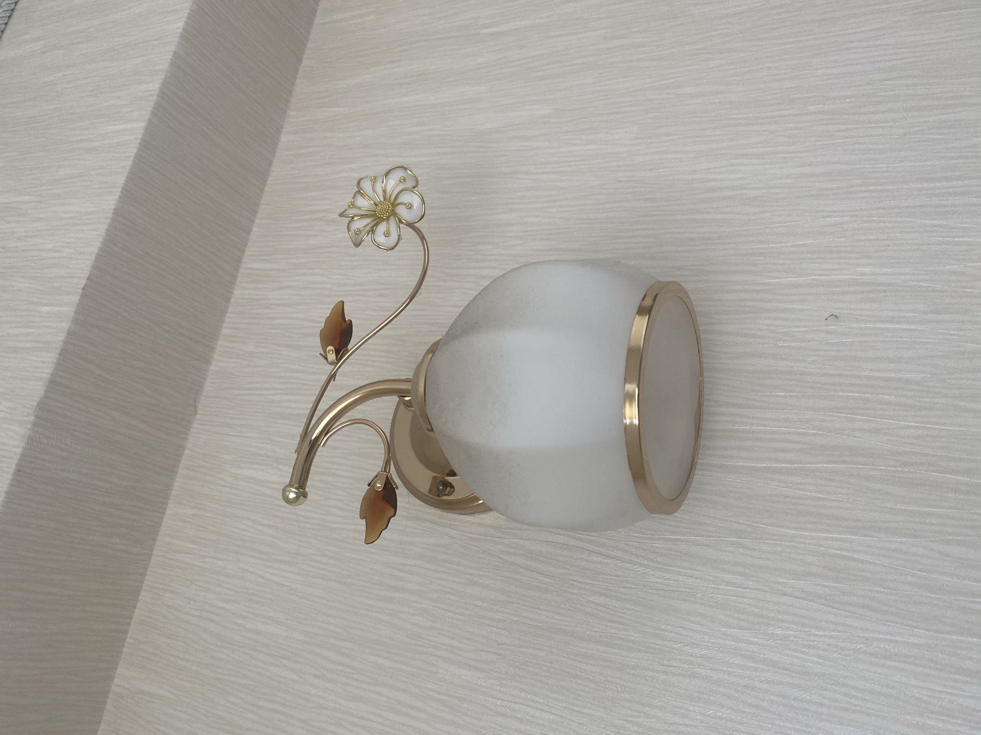 The image shows a close-up view of a wall-mounted light fixture. The fixture has a gold-colored arm extending from a circular base attached to the wall. The arm is adorned with two small, brown, leaf-shaped accents and terminates in a decorative element resembling a flower with white petals and a gold center. The light itself is housed in a frosted white glass shade with a gold band at its base. The wall behind the fixture has a textured, light-colored wallpaper. The lighting suggests an indoor setting, likely a room or hallway. There are no people or actions depicted in the image. The image does not provide any information about its location in Chișinău, Moldova.