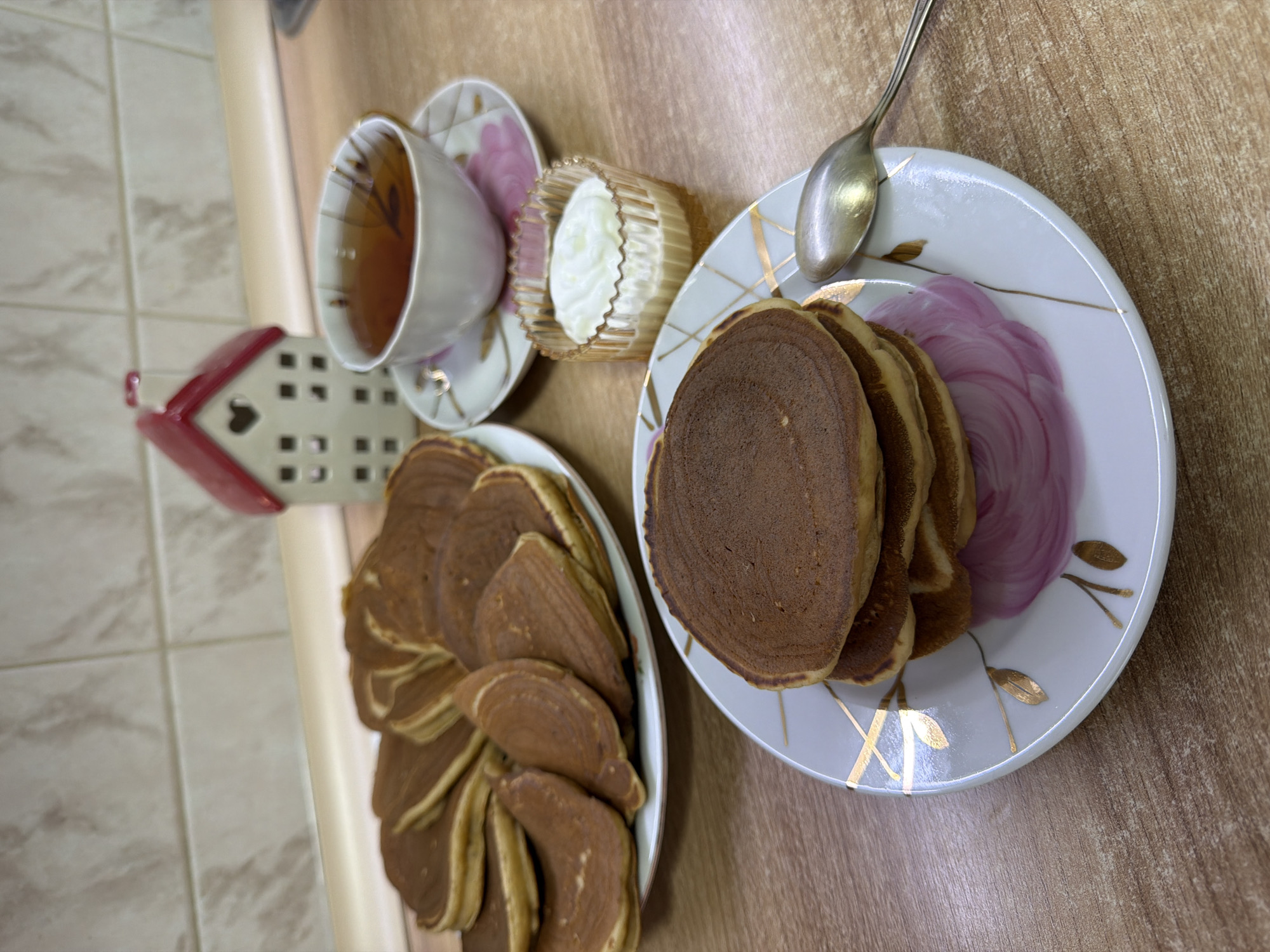 On a wooden countertop in Izhevsk, Russia, a still life is set with breakfast items. In the foreground, a stack of pancakes sits on a white plate adorned with gold leaf and pink floral patterns. A silver spoon rests to the right of the plate. To the left and slightly behind, another plate holds a larger stack of pancakes. A white teacup with a pink floral design sits on its saucer, filled with dark tea. Next to the teacup, a small, fluted glass bowl contains a dollop of white cream. In the background, a decorative white and red house-shaped object stands on a beige tiled surface. The lighting is even, suggesting an indoor setting.