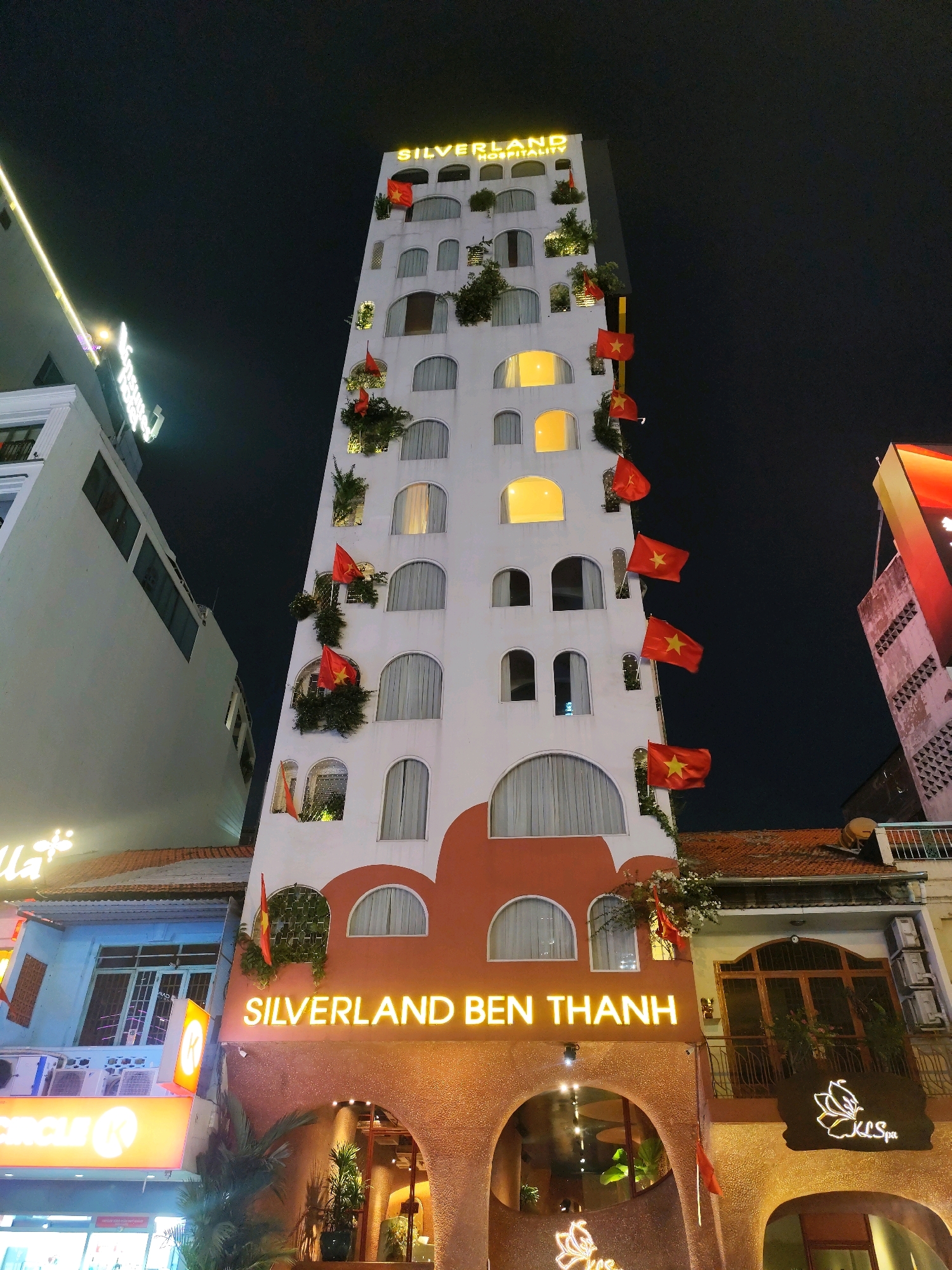 This image captures a night view of Silverland Ben Thanh Hotel, located in Ho Chi Minh City, Vietnam. The hotel is a tall, slender building with a white facade adorned with numerous red Vietnamese flags. The flags are strategically placed along the building's exterior, fluttering in the night air. The hotel features arched windows, some of which are illuminated, casting a warm glow. The name "SILVERLAND BEN THANH" is prominently displayed in neon orange letters at the base of the building. Below the hotel, there are other establishments, including a building with a blue facade and illuminated signage that reads "CIRCLE K". Adjacent to the hotel entrance, a sign for "KL Spa" is visible. The overall scene is a bustling urban environment at night, characterized by the illuminated architecture and commercial signage.