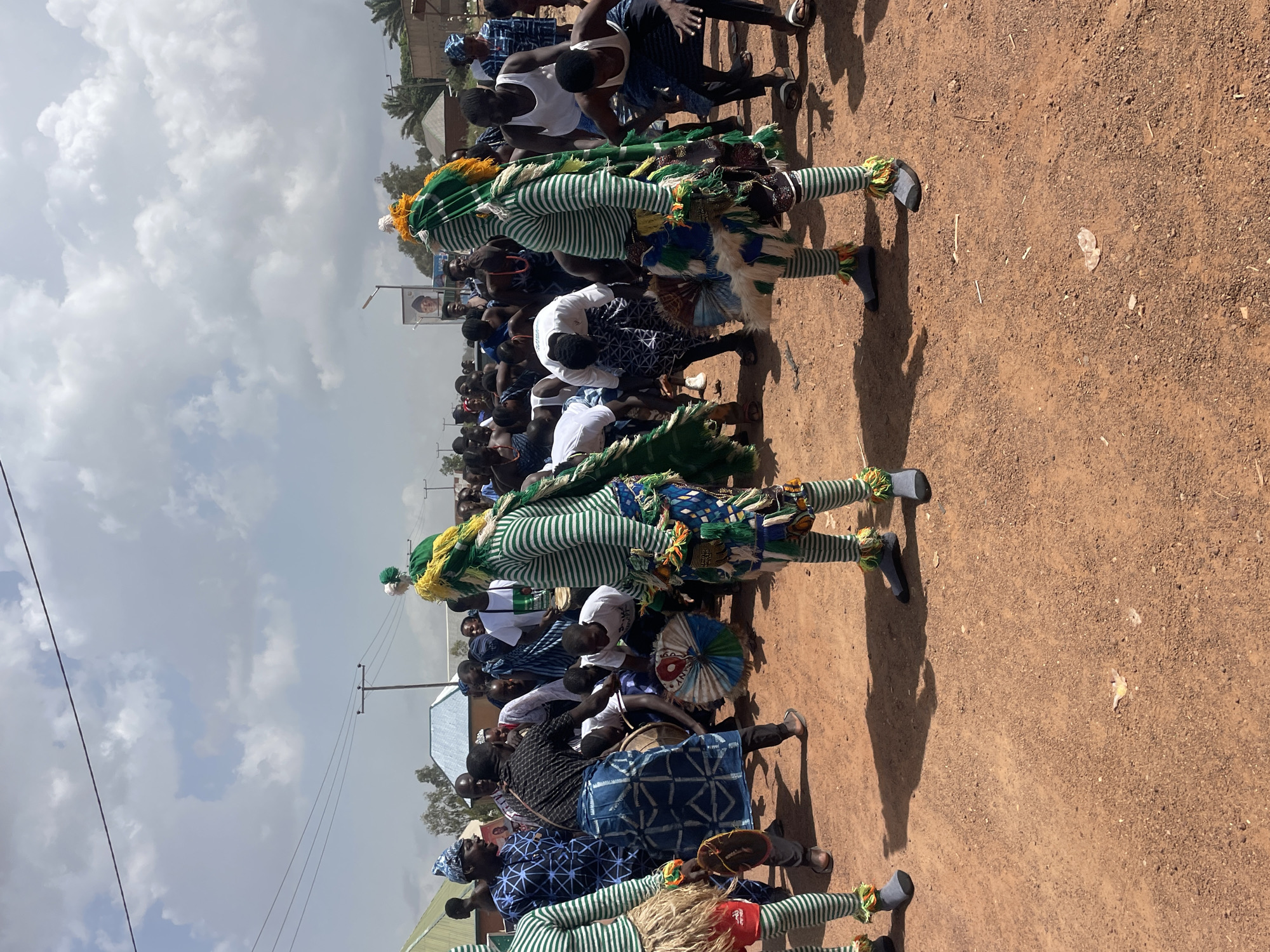 The image shows a public gathering in Durumi, Nigeria. The ground is reddish-brown and dusty. Two figures are prominently dressed in striped green and white costumes with colorful adornments at the head, waist, and ankles. They are surrounded by a crowd of people, many dressed casually. Some are playing drums and other musical instruments. Overhead are power lines against a cloudy sky. A portrait of a man is visible on a pole.