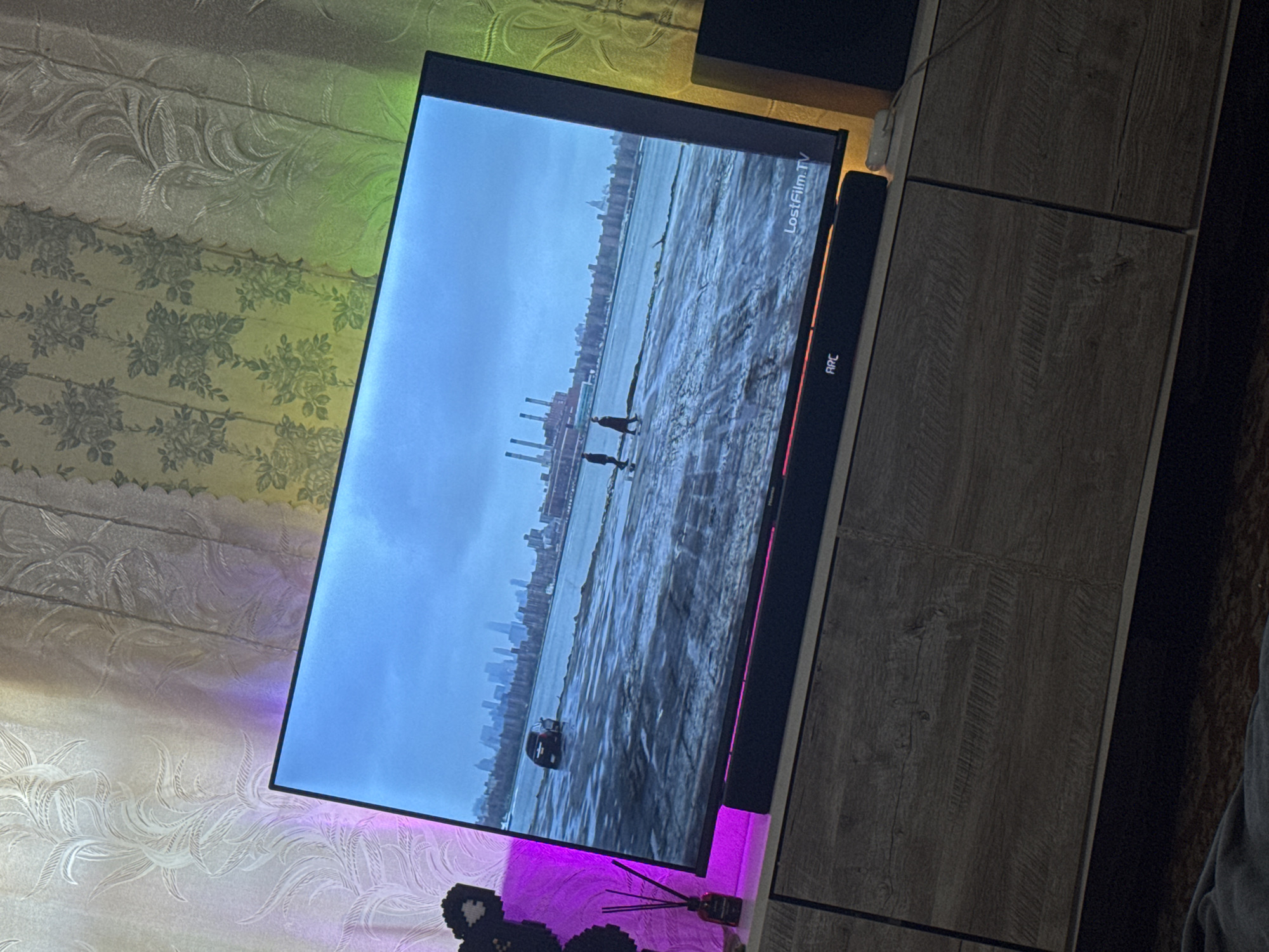 The image depicts an indoor scene in Makiivka, Ukraine, focused on a television displaying a waterfront landscape. The television, a large flat-screen with a visible "Hisense" partial branding and a "LostFilm.TV" watermark on screen, shows a body of water with ice or choppy surface conditions. A dark-colored car is partially submerged near the left bank of the water. Two figures, appearing to be people, walk on the frozen or icy surface towards the right. In the background of the screen, a city skyline with numerous buildings is visible, including an industrial structure with multiple smokestacks on the left and a dome-like building on the right.
Below the television is a black soundbar with the letters "ARC" illuminated, featuring a thin horizontal pink LED strip. The soundbar rests on a light wood-grain television stand with at least two cabinet doors. To the left of the TV, a small dark diffuser bottle with reeds and a black pixelated object (possibly a toy or decoration) are visible. To the right of the TV, a black speaker or subwoofer box stands on the floor, and a white electronic device with a cable is on the TV stand.
The background wall behind the television is covered with patterned wallpaper or curtains, illuminated by colored LED strip lighting. The lighting casts a distinct purple glow on the left, highlighting a light-colored, ornate leafy pattern, and a green-yellow glow on the right, illuminating a darker floral (rose) pattern. A vertical seam separates the two wallpaper sections. The foreground shows a dark, possibly carpeted floor, with a dark blanket or fabric partially visible at the bottom.