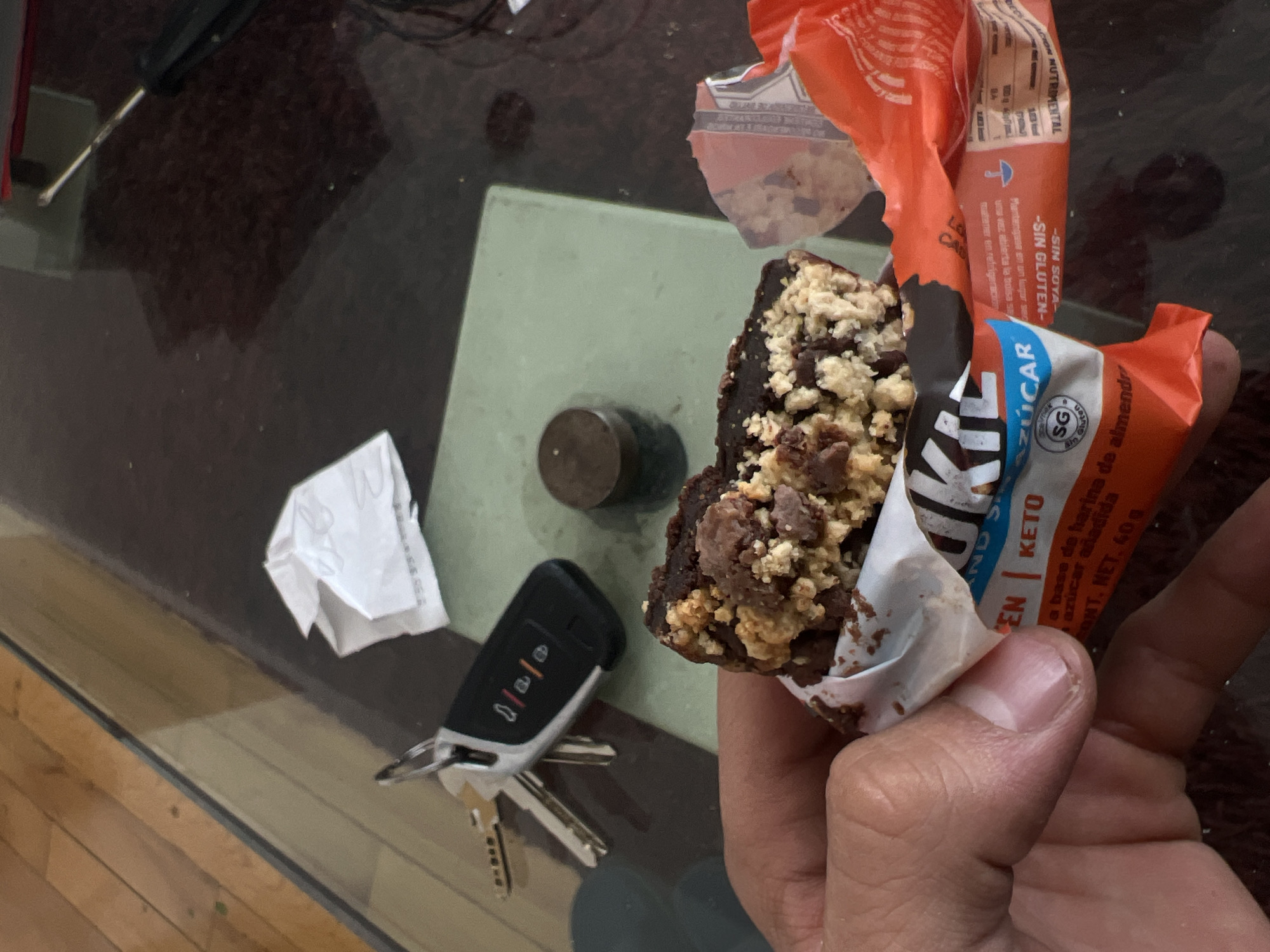 A close-up, eye-level shot shows a hand holding a partially eaten food bar, likely a brownie or chocolate bar with a light-colored crumble topping, on a glass-top table. The bar's orange and white wrapper is peeled back, revealing product information. On the table, to the left of the bar, are a set of car keys with a black modern key fob and two silver keys. Further to the left, a crumpled piece of white paper rests on the table, and a small, dark cylindrical object is also visible. Part of a screwdriver is faintly seen in the top left corner, and the wooden floor is reflected through the glass table.
The food bar wrapper has prominent text in Spanish, including "SIN SOYA-" (Soy-free), "SIN GLUTEN-" (Gluten-free), and "KETO". Other visible phrases indicate it is "a base de harina de almendra" (almond flour based) and contains "azúcar añadida" (added sugar). The packaging also mentions "CONT. NET. 40 g" (Net Content: 40g) and instructions to "Manténgase en un lugar fresco y seco" (Keep in a cool and dry place) and "mantener en refrigeración una vez abierta la bolsa" (keep refrigerated once the bag is open). A "SG" logo confirms its "Sin Gluten" status. The scene depicts a casual indoor setting, possibly at a home or office in Mexico City, given the Spanish language on the product packaging. The activity is the consumption of a snack bar.