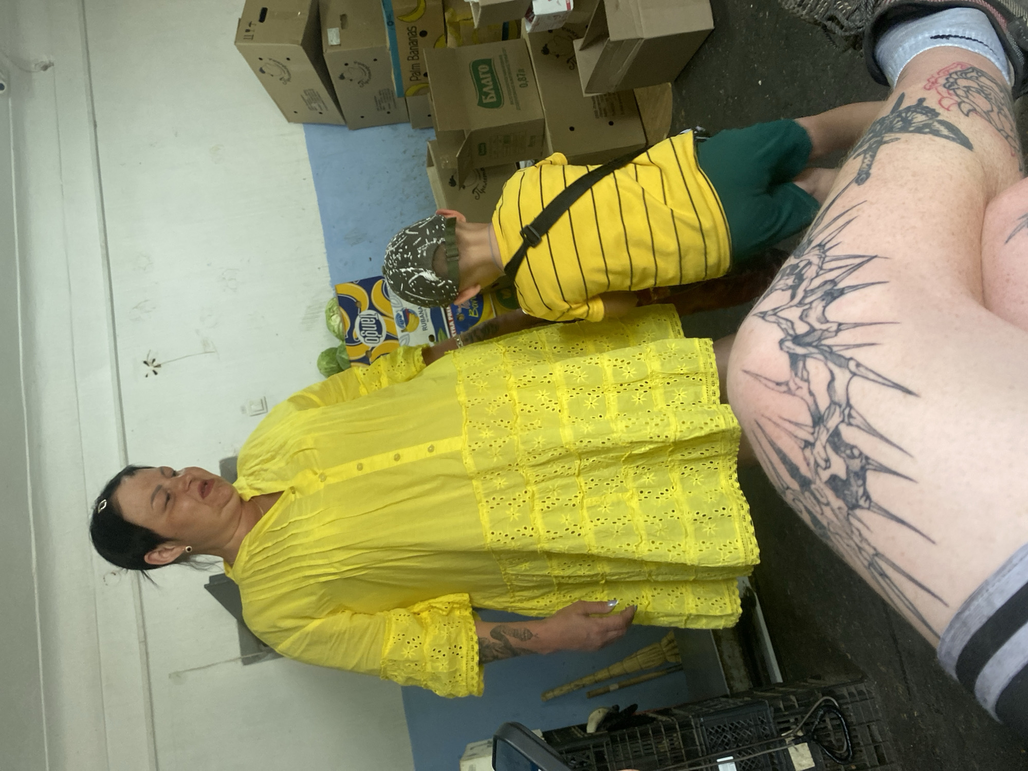 In a dimly lit interior space, a woman in a bright yellow dress stands looking down with a distorted expression. To her right, a young boy in a yellow striped shirt and green shorts faces away, wearing a patterned cap. The background consists of white and blue walls stacked with cardboard boxes, some labeled with text and graphics including "Palm Bananas" and "Благо". The foreground shows a person's leg with extensive black tattoos, including a large, intricate design on the thigh. A black tote bag strap is visible across the boy's chest. The floor is concrete, and various items, including a black crate and brooms, are present in the scene.
