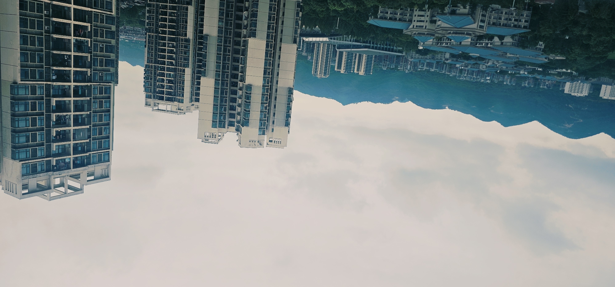 The image captures a wide view of a residential area in the city of Chongqing High-tech Zone, China, under an overcast sky. In the foreground and midground, several multi-story apartment buildings are visible. The buildings vary in design and height, with some featuring a light-colored facade and numerous windows. In the background, a range of lush green mountains forms a backdrop to the urban landscape. The overall scene depicts a dense urban development nestled amongst natural topography, with the city extending towards the foothills of the mountains. There are no people or vehicles clearly discernible in the image.