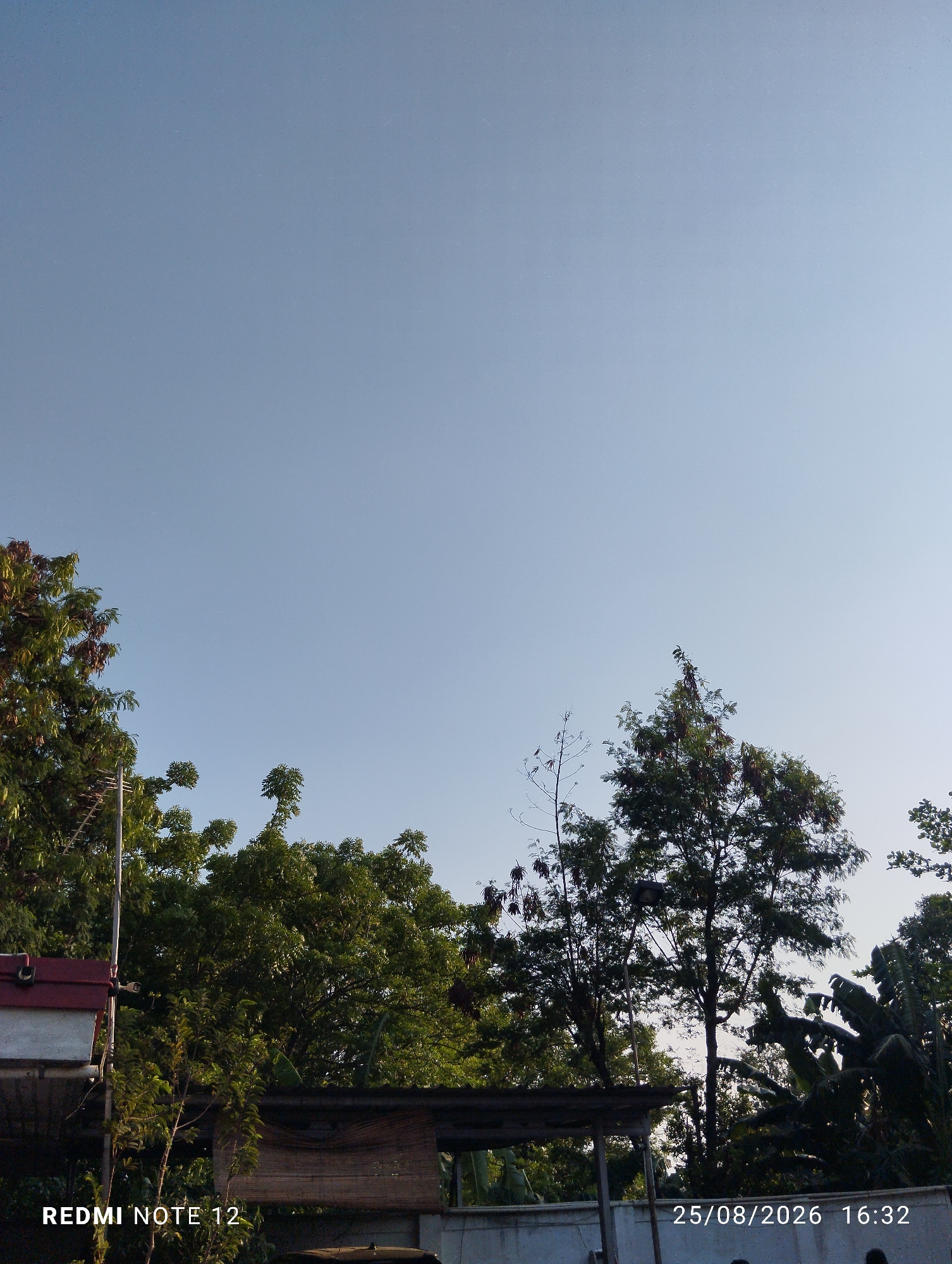 A daytime, eye-level, outdoor shot captures a clear blue sky above a densely wooded area. In the foreground, the top of a white and red structure is visible on the left, with a metal antenna extending upwards. Trees with lush green foliage fill the middle ground, and to the right, banana trees are present. The bottom of the image displays a white wall with a black car parked in front of it. Text overlays at the bottom left read "REDMI NOTE 12", and at the bottom right, "25/08/2026 16:32". The scene appears to be in a residential area of Bekasi, Indonesia.