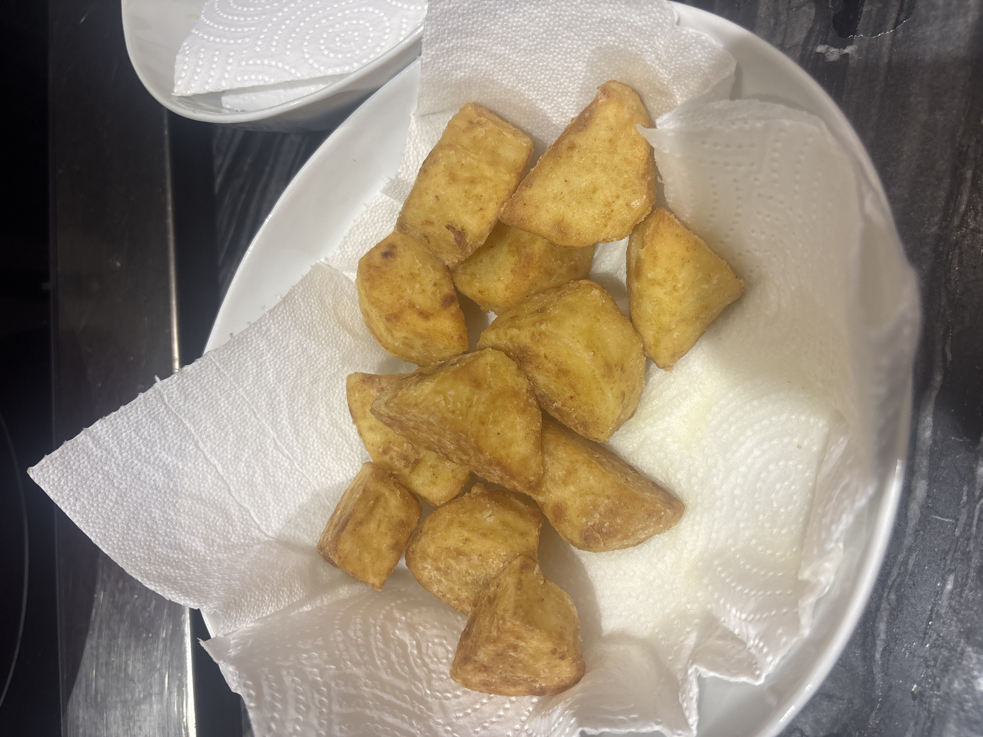 A close-up, top-down shot shows a pile of golden-brown, fried potato wedges resting on white paper towels. The paper towels are placed inside a white ceramic bowl, which is on a dark, speckled countertop. In the background, a stainless steel appliance is visible, possibly a stovetop or a part of a kitchen counter. The lighting is bright, highlighting the texture and slight crispiness of the potatoes. There are no people or any indication of location in Dubai, United Arab Emirates.