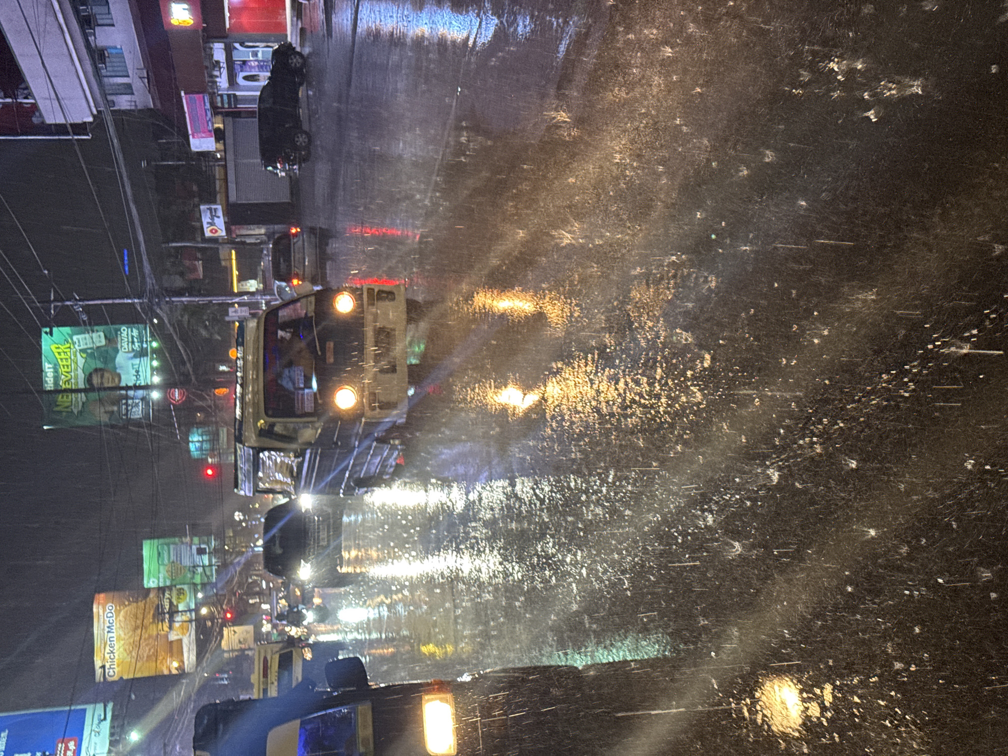 The image depicts a street scene in Davao City, Philippines, at night during a heavy downpour. The wet asphalt road reflects the neon lights of billboards and streetlights, creating a shimmering, blurred effect. Several vehicles are visible, including a blue jeepney on the left with its headlights on, a black SUV in the middle also with its headlights illuminated, and a yellow passenger van with its hazard lights flashing. A white tricycle with its headlights on is also present. The rain is falling heavily, with visible streaks and splashes on the road and windshields. Billboards advertising "Chicken McDo" and other businesses are illuminated in the background. Power lines crisscross the sky, adding to the urban feel of the scene. The overall impression is one of a bustling city street transformed by the intensity of the rain.