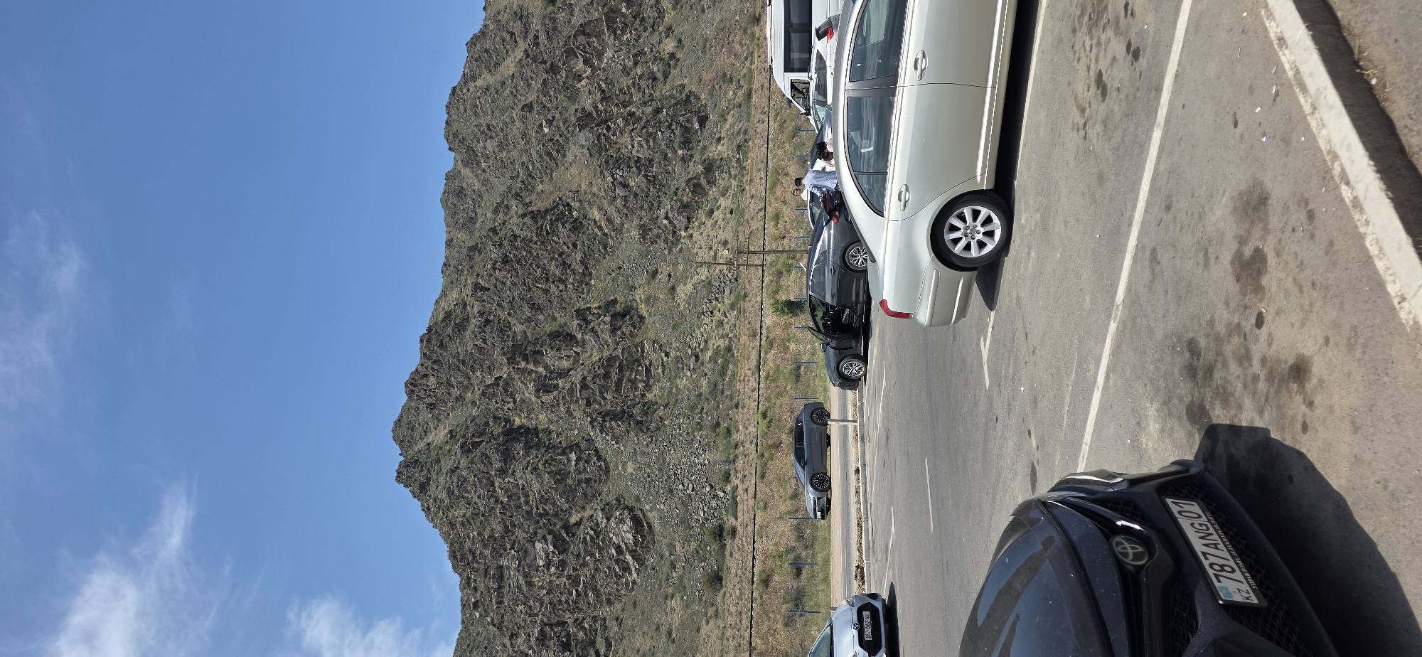 The image depicts a parking lot scene with a mountainous backdrop. In the foreground, a black Toyota sedan is parked, its license plate reading "KZ 787 ANG 01". To the right of the black sedan, a silver sedan is parked. Further back, a dark grey sedan with its driver's door open is parked next to a light-colored sedan. A white van is visible in the background. Several individuals are standing near the dark grey sedan. The environment features a paved parking lot, a road, a grassy area with a fence, and a rugged, rocky mountain range under a clear blue sky. The location is identified as Sasyk-Bulak, Kyrgyzstan.
