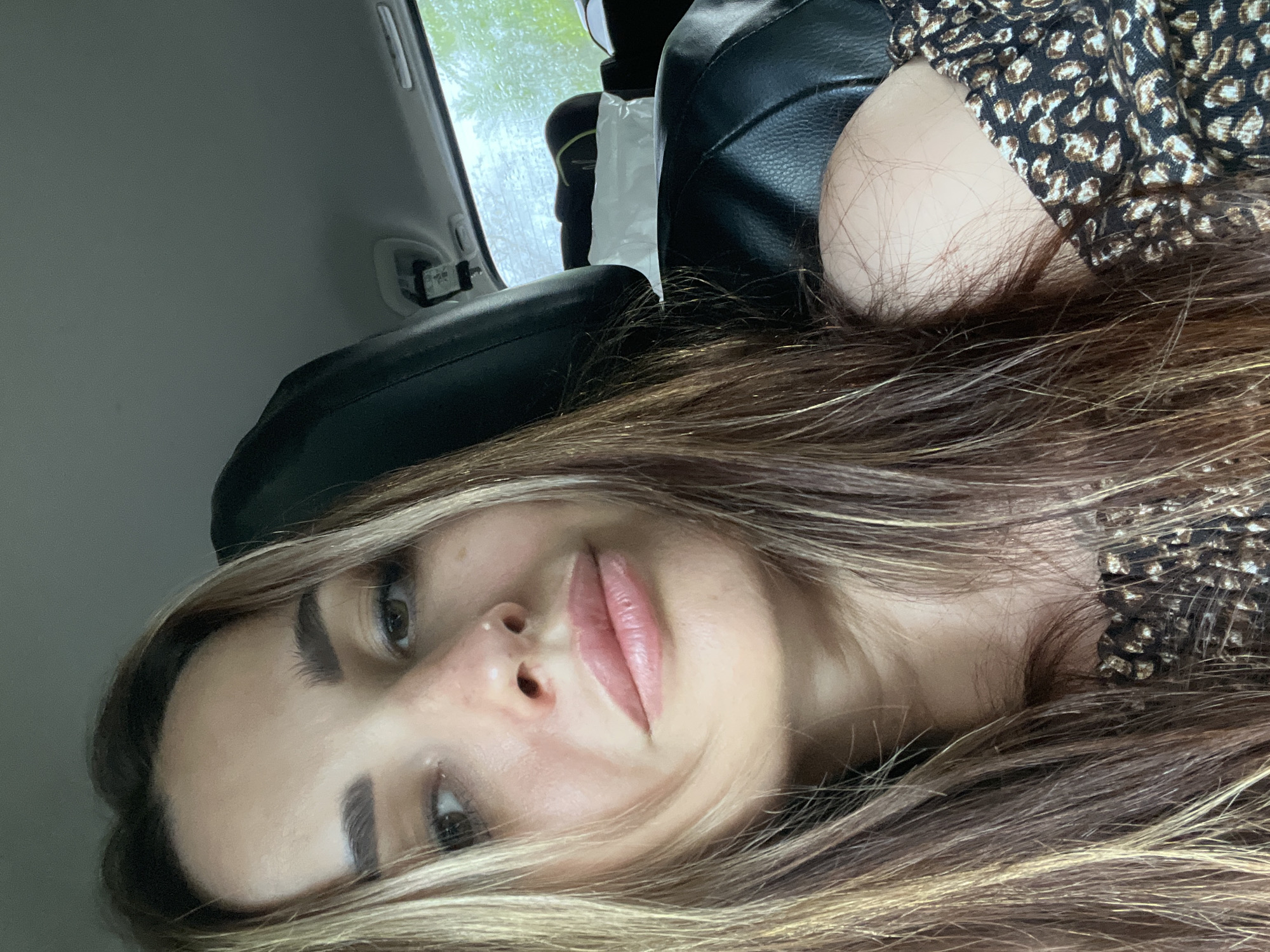 A medium shot shows a woman in the driver's seat of a car, looking towards the camera. She has long, brown hair with blonde highlights, styled to fall around her face and shoulders. She is wearing an off-the-shoulder top with a leopard print pattern. Her lips are full and appear to be glossed. Her eyes are brown, and her eyebrows are well-defined. The background shows the interior of the car, including a black leather headrest and a glimpse of the rear window, which appears to be slightly wet, suggesting it might be raining outside. There are also some bags or items in the back seat. The lighting is soft and natural.