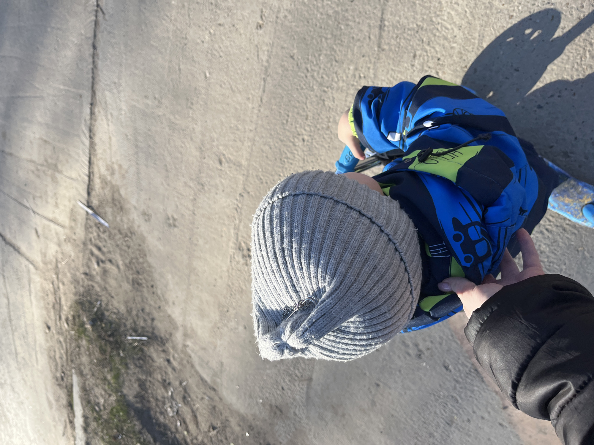 A top-down shot shows a child in a grey knit cap and a blue and black jacket. The child appears to be on a blue scooter. An adult's hand in a black jacket sleeve reaches towards the child. The scene is set on a concrete surface. There is a discarded cigarette and debris visible on the concrete surface in the background. The image doesn't provide the exact coordinates to locate it in Chaikovsky. The location can't be determined from the image.