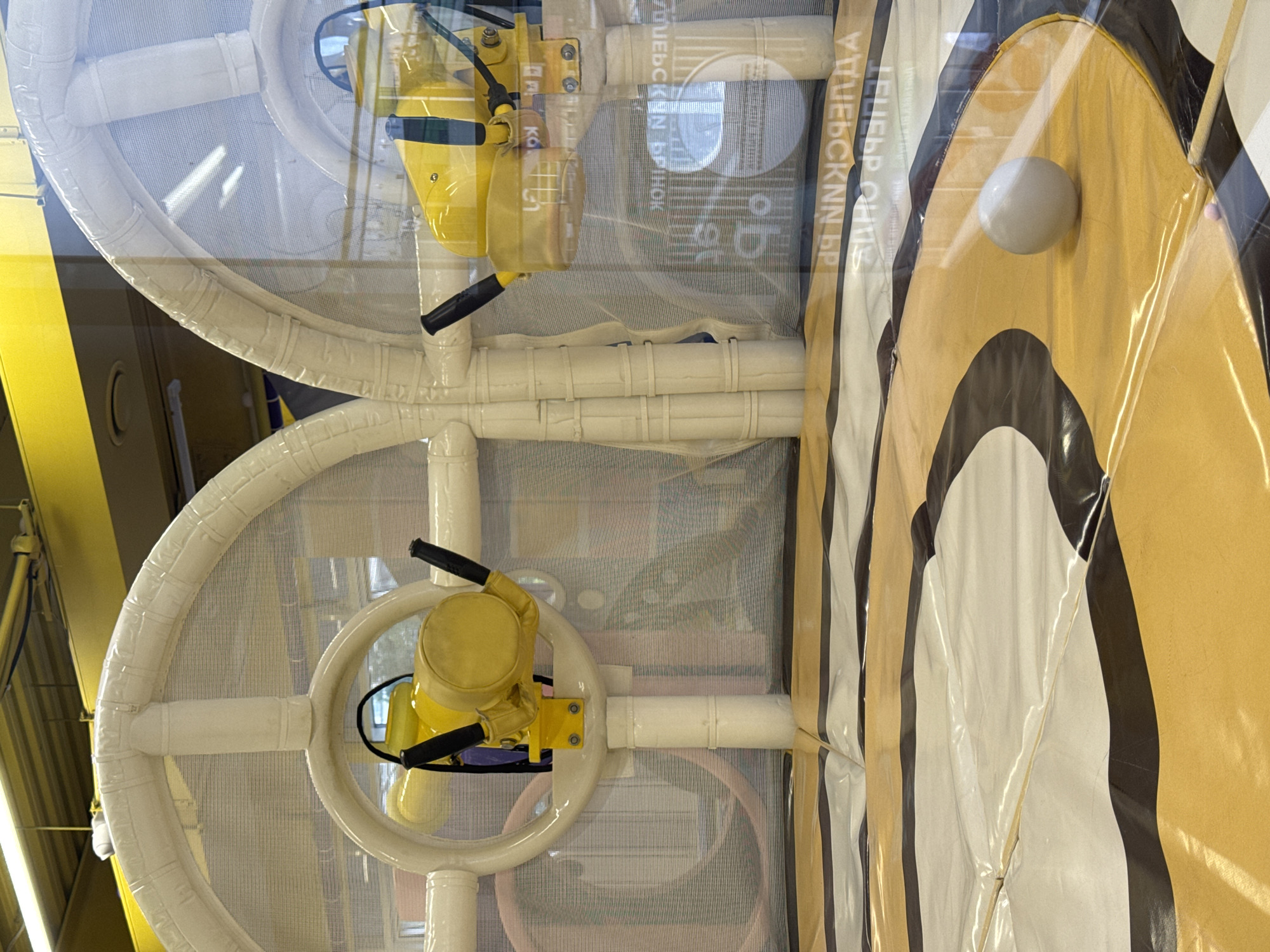 The image shows a children's play area, likely indoors, featuring playground equipment. There are two large, circular structures made of white tubing, resembling steering wheels or control panels, each with a yellow apparatus attached. The apparatus have black handles. The background includes a white mesh or netting behind the circular structures. To the right, there is a striped pattern in shades of yellow, white, and black. The overall impression is of a play area designed for children. The location is not evident.