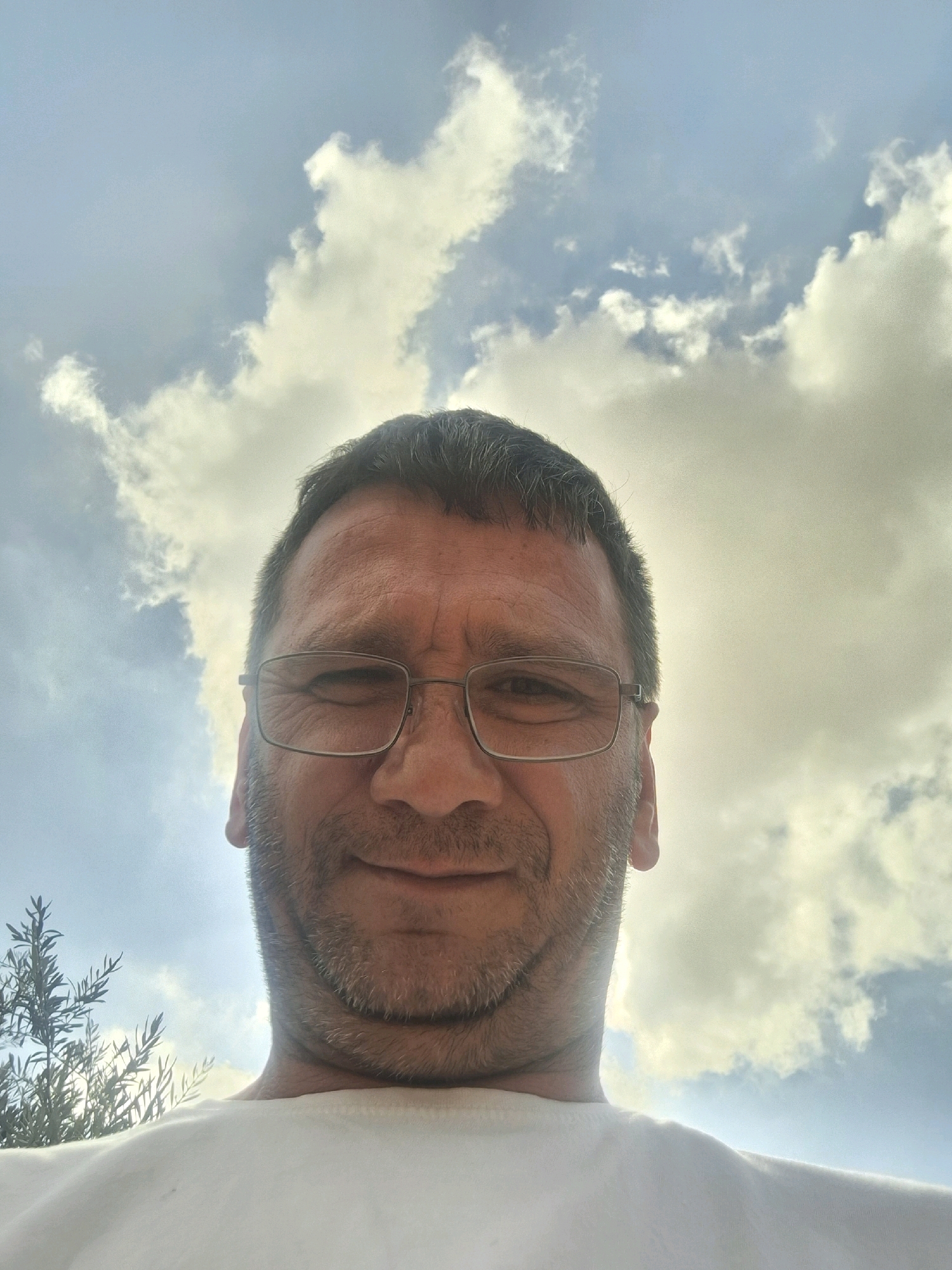 The image is a low-angle, close-up selfie taken outdoors against a bright, cloudy sky. The subject is a middle-aged man with short, dark hair, glasses, and stubble. He is wearing a white t-shirt. The background includes a tree with fine leaves on the left side of the frame and fluffy white and gray clouds against a light blue sky. The lighting suggests it is daytime, possibly late afternoon, with the sun partially obscured by clouds. The camera appears to be held above and in front of the man, looking up at him. The overall impression is a casual, personal photograph taken in an outdoor setting. There is no specific identifiable location within the city of Kiryat Gat, Israel mentioned or visually represented in this image.