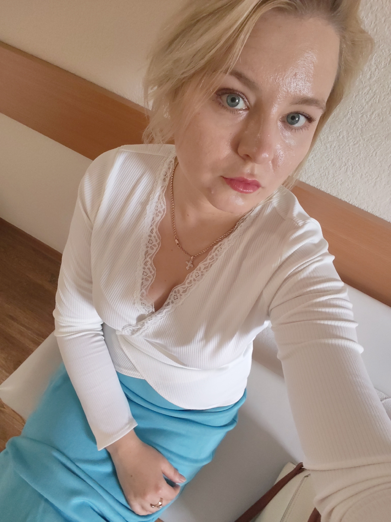 In an indoor setting with a textured, off-white wall and a wooden beam, a fair-skinned woman with blonde hair and blue eyes is captured from a high angle, looking towards the camera. She is wearing a white, ribbed long-sleeved top with a lace trim neckline and a bright blue skirt. A gold chain with a cross pendant is visible around her neck. Her right hand rests on her lap, adorned with a ring. The lighting suggests an ambient light source. The image does not provide enough visual information to determine the specific location within Izhevsk, Russia.