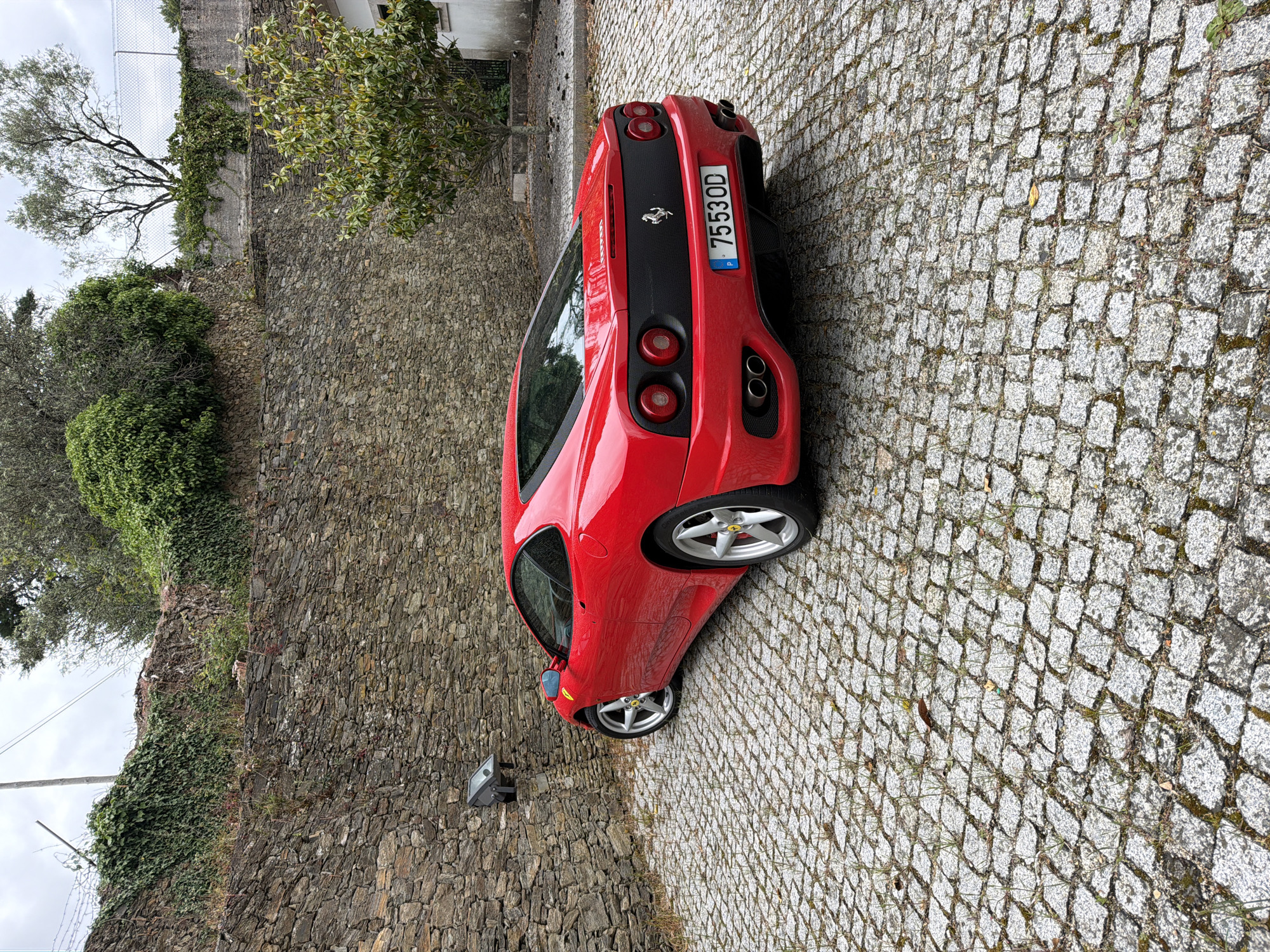 A red Ferrari sports car is parked on a cobblestone road next to a stone wall covered with ivy and greenery. The car's license plate number is 7553OD, and it has a Portuguese registration sticker. The scene is in Vila Real, Portugal, judging by the license plate. The weather appears overcast. A streetlight fixture is mounted on the stone wall. A tree is visible above the wall.