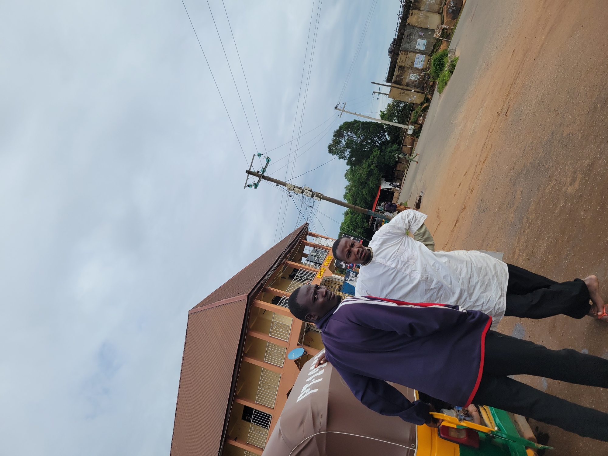In the city of Jos, Nigeria, two men stand near a yellow and green three-wheeled auto-rickshaw, commonly known as a "keke". One man, wearing a navy blue jacket with red and white trim, faces the camera with his left hand on the vehicle. The other man, dressed in a white traditional outfit, stands slightly behind him, also looking towards the camera. The background features a multi-story building with a brown roof and balconies, a power pole with multiple wires, and a street with a dirt shoulder. The sky is overcast, indicating a cloudy day.