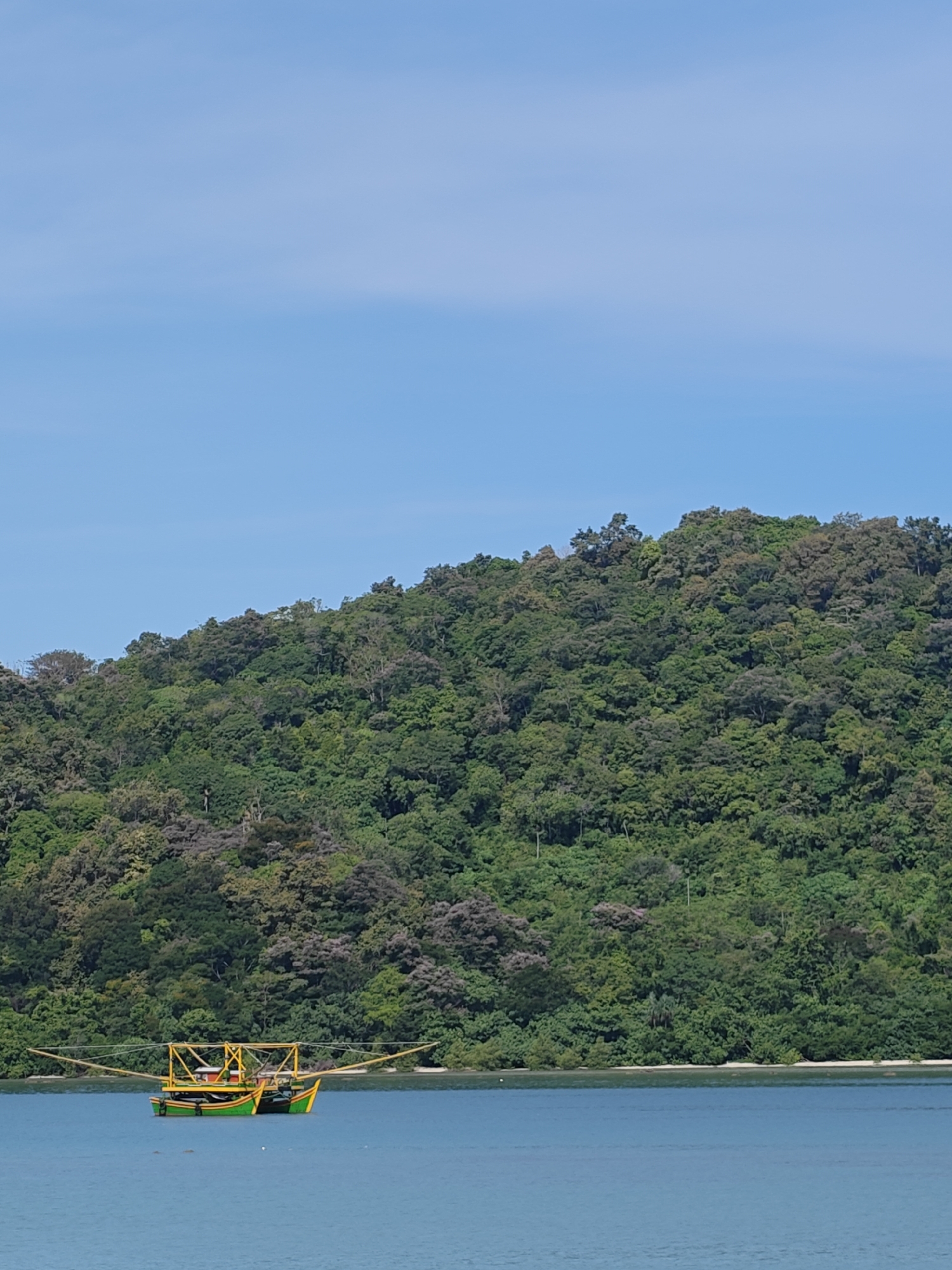 The image depicts a traditional Indonesian fishing boat in the water off the coast of Layeun, Indonesia. The boat has a distinctive configuration, with a green and yellow hull and outrigger arms extending from either side. It is located near a densely vegetated island with a blue sky above. The water is calm and blue, reflecting the sky.