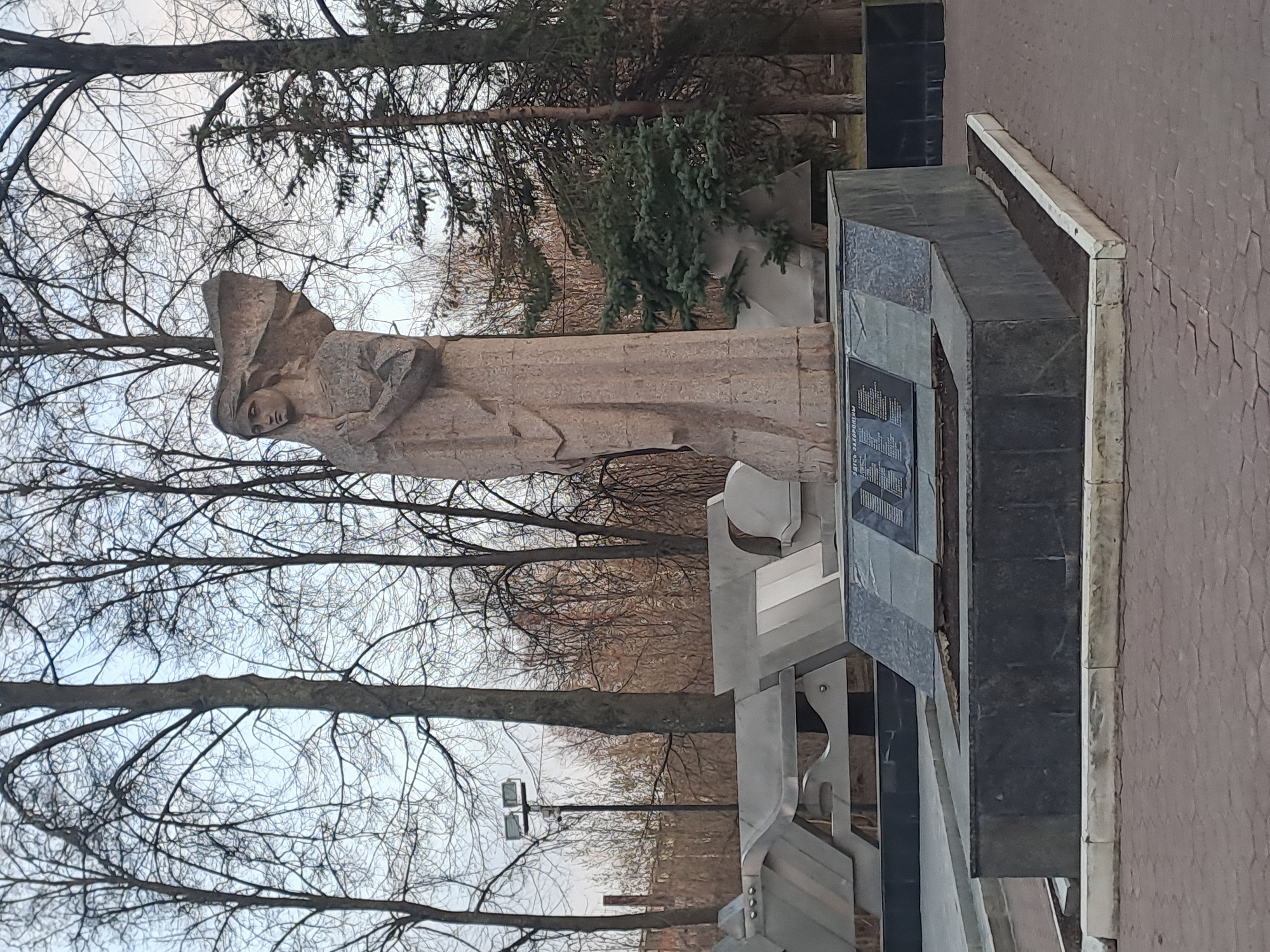 The image shows a monument in Elektrostal, Russia. The monument consists of a stone sculpture of a woman and a granite structure with inscriptions. The woman is depicted in a flowing garment, with her head covered and arms crossed. The granite structure features rectangular plates with text written in Russian. Trees and bare branches are visible in the background, suggesting it may be late autumn or winter. A light pole and paved walkway are also part of the scene.
