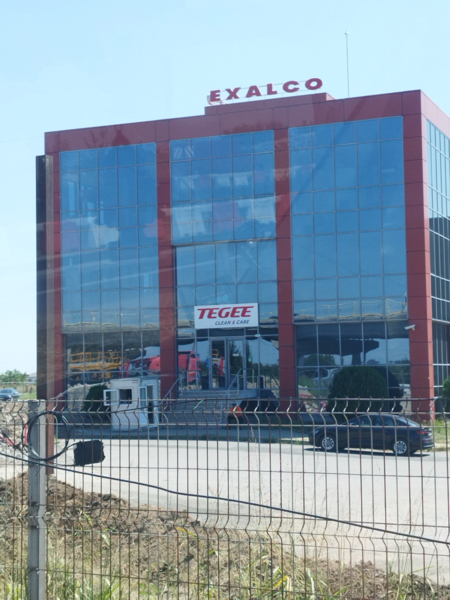 The image depicts the exterior of a modern, multi-story building with a red facade and extensive blue glass windows. The building is topped with the red lettering "EXALCO". Below the "EXALCO" sign, a smaller red rectangular sign reads "TEGEE CLEAN & CARE". In front of the building, a chain-link fence runs across the foreground, partially obscuring the view of the building's base and the surrounding area. Behind the fence, there are piles of dirt and dry grass, suggesting an outdoor or construction environment. A dark-colored sedan is parked on a paved road in front of the fence and to the right of the building. In the reflection of the building's windows, yellow construction equipment and red vehicles are visible, indicating activity or storage of machinery behind the building. The sky is clear and blue. The building is located in the city of Bragadiru, Romania.