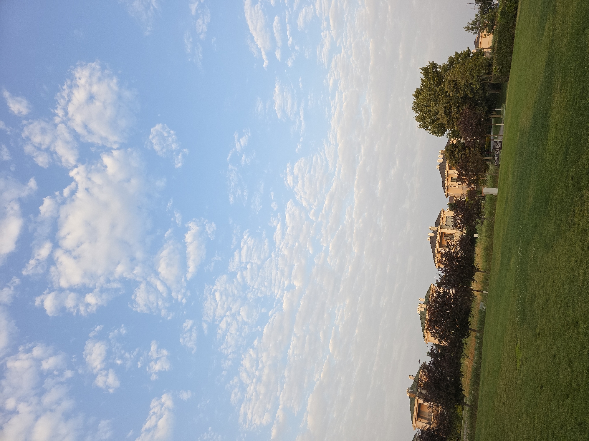 This is a photograph of a residential area in Surakhany Raion, Azerbaijan. The image features a clear blue sky with scattered white clouds, and a neatly mown green lawn in the foreground. In the middle ground, there is a row of multi-story houses with tiled roofs, interspersed with trees. The houses appear to be modern suburban residences. The overall impression is one of a peaceful, well-maintained suburban setting. There are no people or animals visible in the image.
