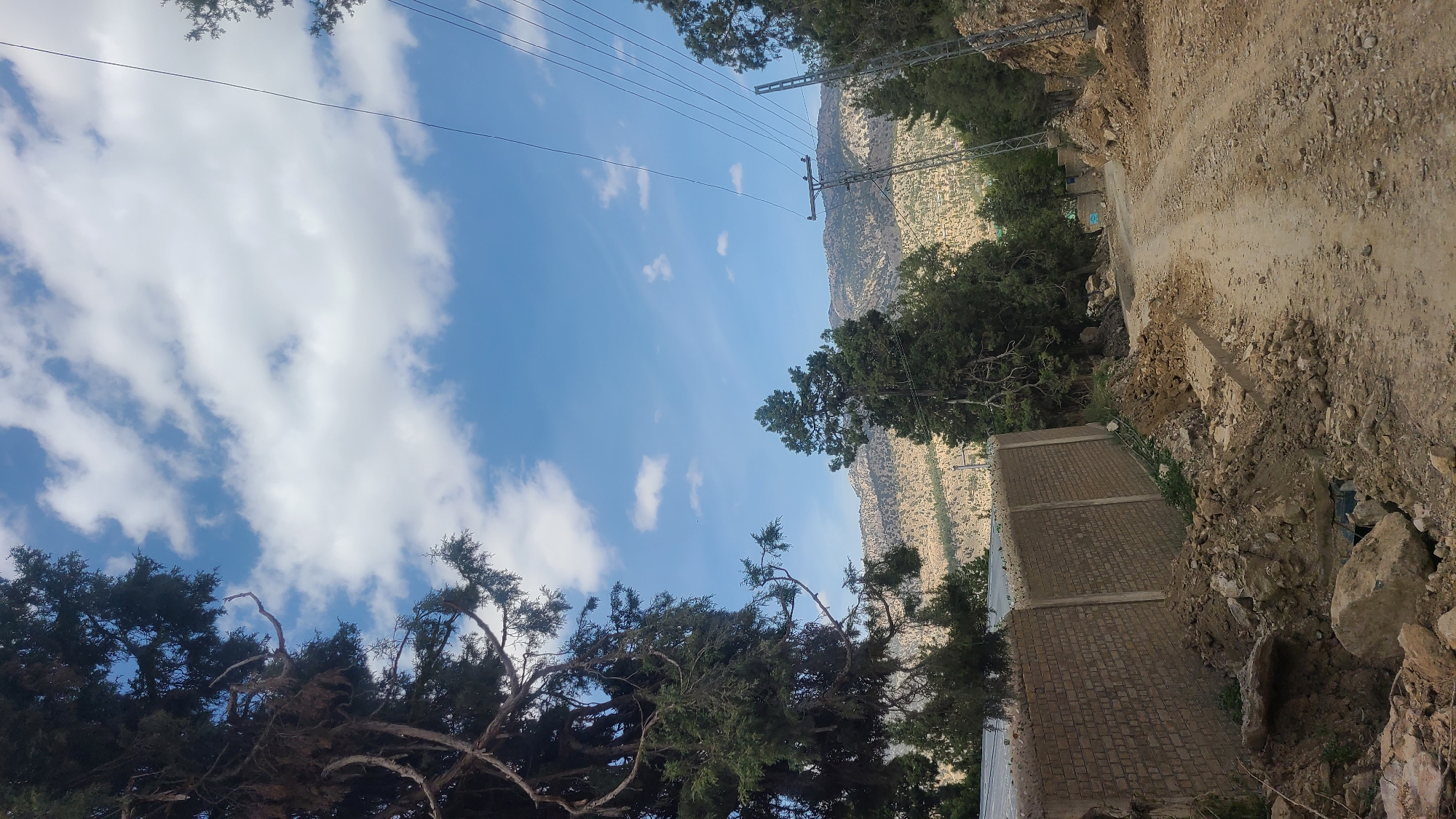 The image depicts a landscape in Ziarat, Pakistan, featuring a dirt road winding along the side of a mountain. To the left, a brick structure is visible, potentially part of a building or wall. The sky above is blue with scattered white clouds, and power lines stretch across the scene. Trees are present along the roadside and in the foreground, adding greenery to the otherwise arid environment. The overall impression is of a rural, mountainous region.