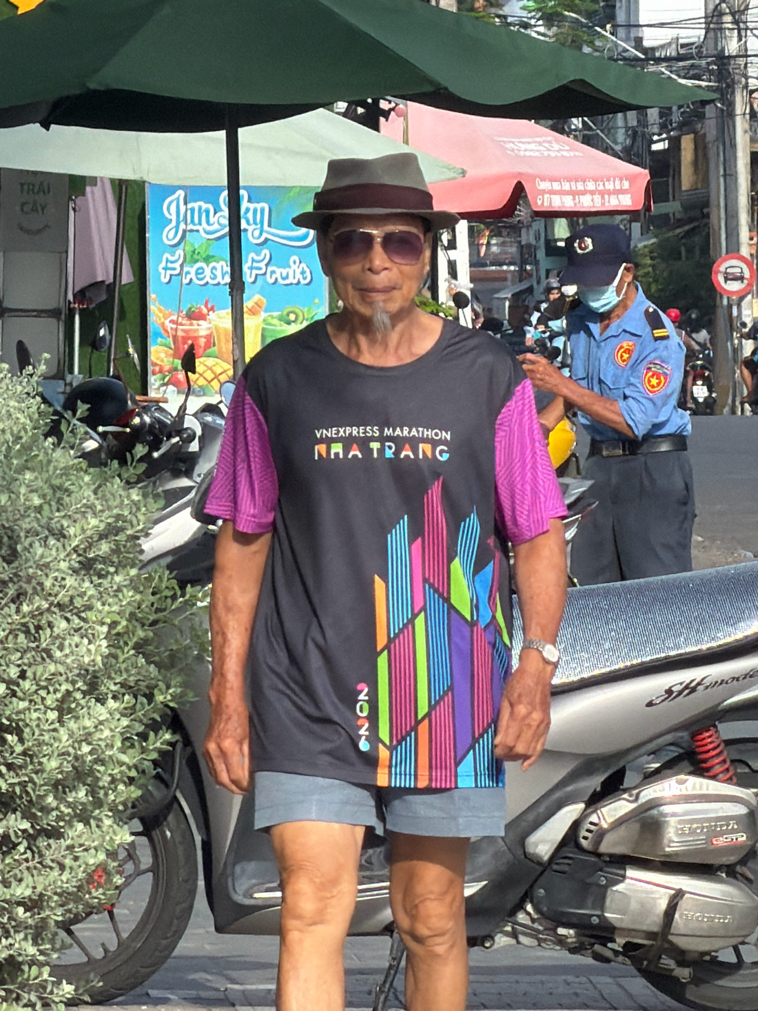 In Nha Trang, Vietnam, an elderly Vietnamese man with sunglasses and a hat stands prominently in the foreground, facing the camera. He wears a black t-shirt with purple sleeves featuring a colorful graphic print and "VNEXPRESS MARATHON NHA TRANG 2026" text. He is also wearing light blue shorts and a watch on his left wrist. Behind him, a uniformed security guard in a blue shirt and cap, wearing a face mask, is partially visible, appearing to adjust his uniform. The background includes a juice stand with colorful signage, several parked motorcycles, a green umbrella, and a red awning. The scene takes place on a sunny day, likely on a street in the city.