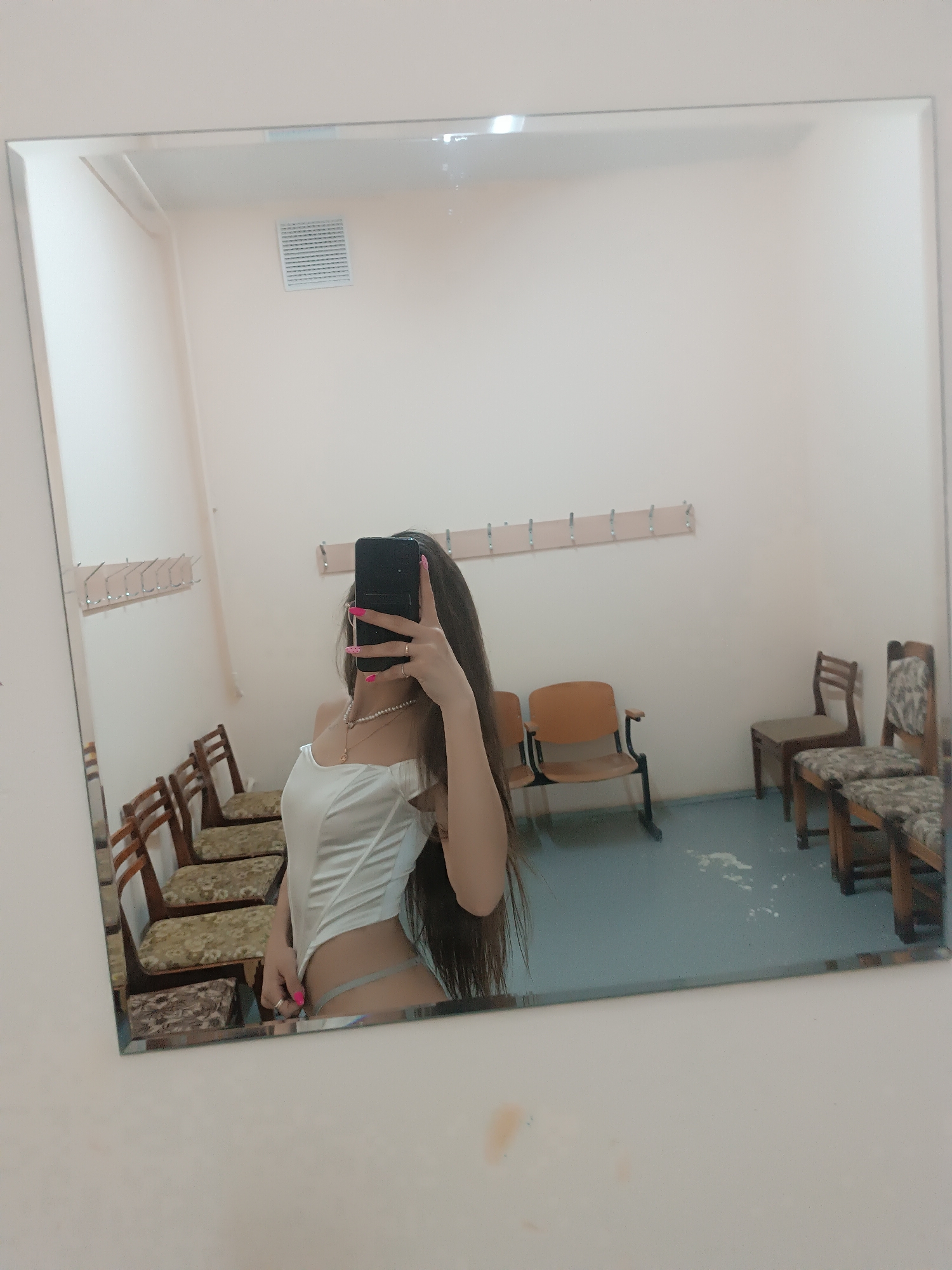 A photo shows a reflection in a mirror. The reflection shows a woman in a white top and a light gray thong. She is holding a smartphone to take a selfie, obscuring her face. The background appears to be a changing room with several chairs and coat hooks on the wall. The floor is blue-gray. The city and state of Unknown, Unknown, are not confirmed by the photograph.