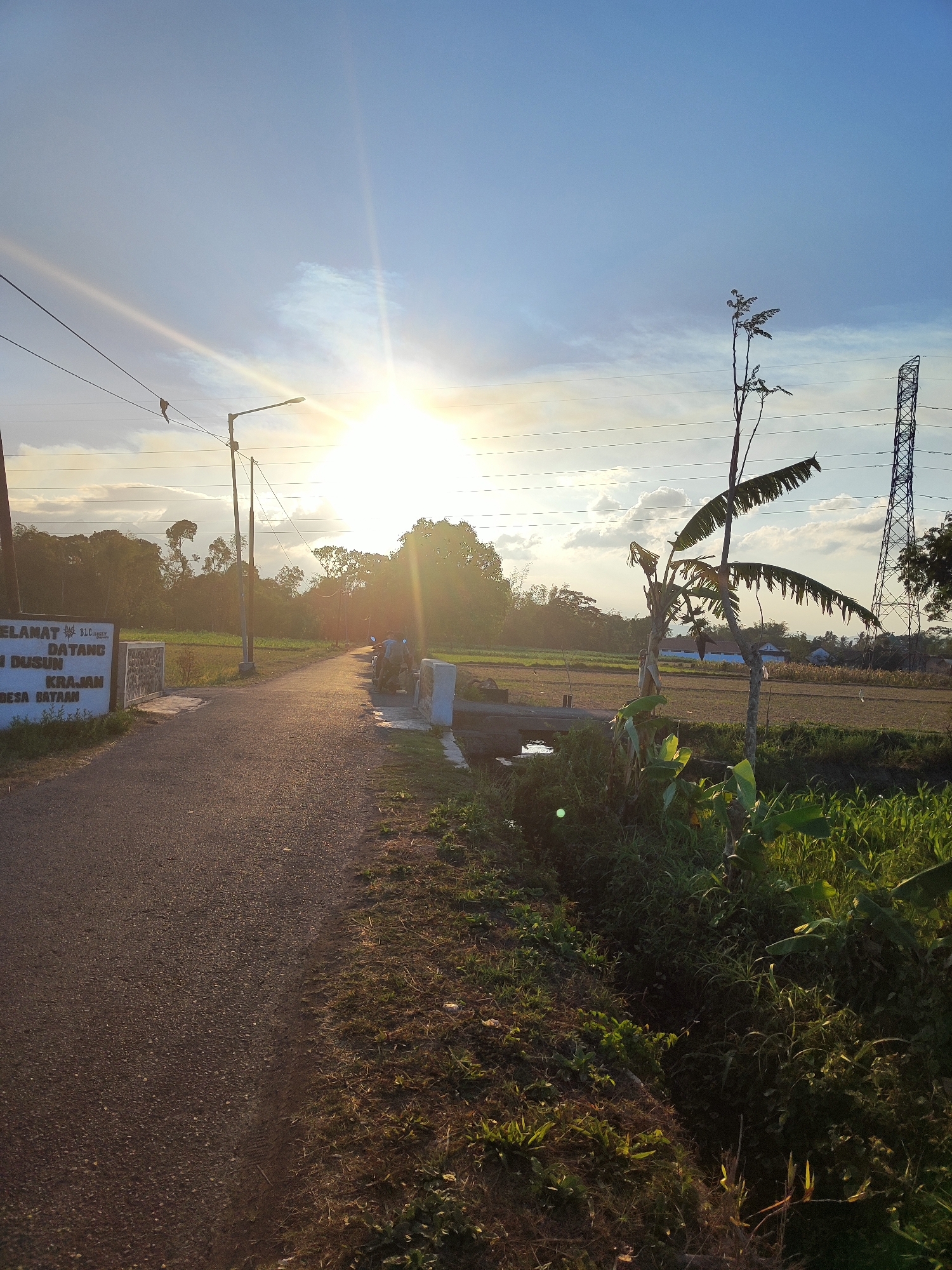 The image depicts a rural road at sunset in Bondowoso, Indonesia. The sun is low on the horizon, casting a warm glow and lens flare across the scene. On the left side of the road, a white sign with text is partially visible, with the words "SELAMAT DATANG DI DUSUN KRAJAN DESA BATAA" clearly legible. A street light stands tall next to the sign. The road itself is asphalt and curves slightly into the distance. On the right side of the road, a small channel of water runs parallel to the road, bordered by lush green vegetation. A banana tree with sparse leaves stands prominently in the foreground on the right. In the middle ground, a bridge crosses the channel, and two individuals on a motorcycle are visible on the bridge. Beyond the immediate roadside, a field stretches out, with some agricultural structures and houses in the distance. Power lines are visible crisscrossing the sky, and a tall transmission tower is present on the far right. The overall atmosphere is serene and tranquil, characteristic of a rural Indonesian sunset.