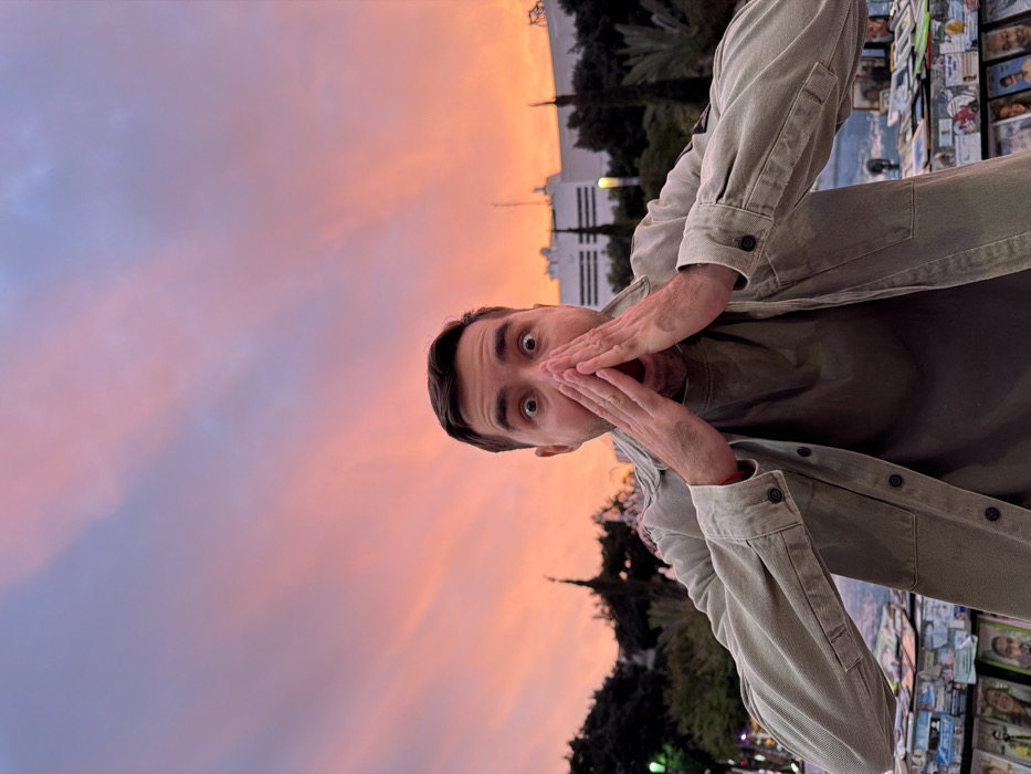 A man, seen from a low angle, stands with a surprised expression, his hands framing his mouth. He is wearing a casual outfit: a muted green t-shirt and a light green jacket. In the background, a striking sunset paints the sky in hues of pink and orange. Beyond the man, trees and a building are visible against the vibrant sky. Adjacent to him, a display of printed materials adds context to the scene.