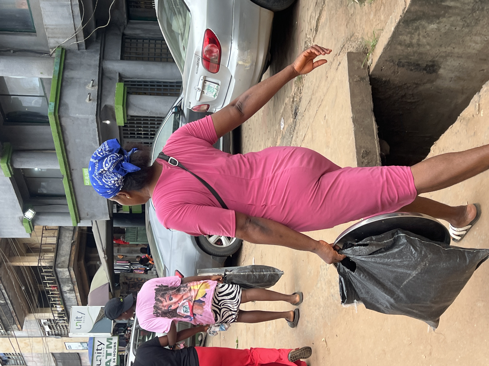 In Nkpor, Nigeria, a woman in a bright pink dress and a blue bandana walks down a dirt street, carrying a large black trash bag and a metal tray. The street is lined with buildings, including a Unity Bank. Cars are parked along the street. The woman appears to be heading towards a covered market area, where other people are visible. The scene suggests a busy, commercial area with everyday life unfolding.