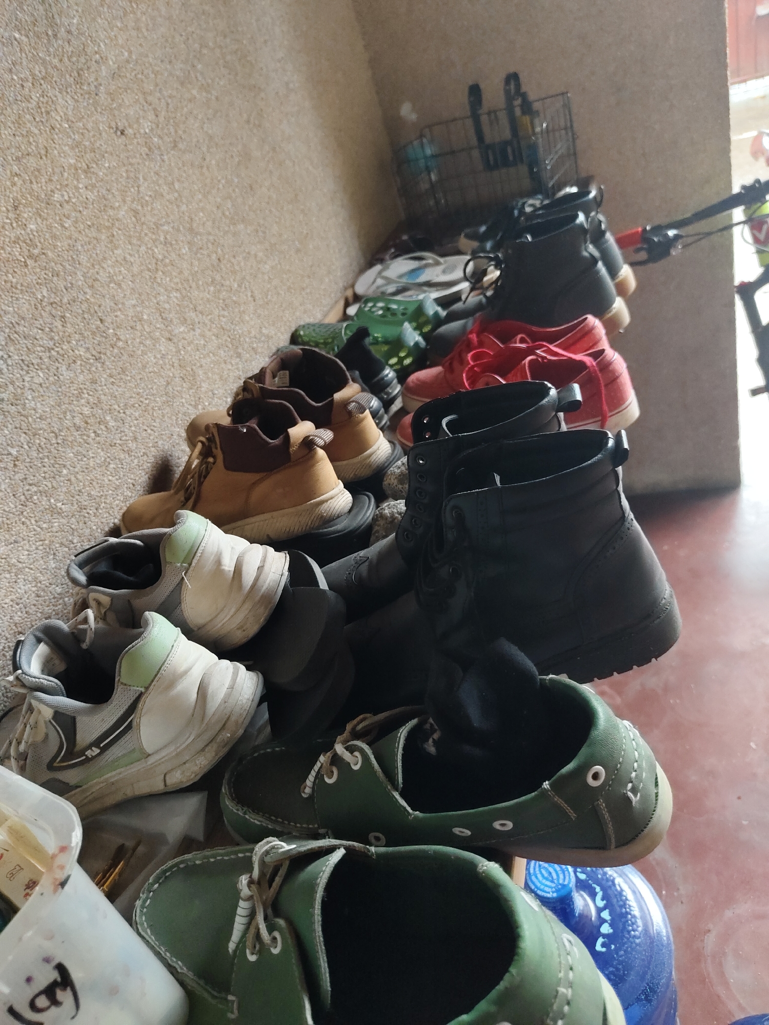 The image shows a collection of shoes and boots arranged on a floor against a textured wall. The items include several pairs of athletic shoes, work boots, and casual shoes, mostly in neutral colors such as beige, black, white, and green, with a pair of red shoes also visible. In the background, a bicycle with a basket is partially visible, leaning against the wall. The setting appears to be an outdoor or semi-outdoor area, possibly a porch or entryway, with a concrete floor. The image does not provide any specific visual cues that can definitively identify the location as Davao City, Philippines.