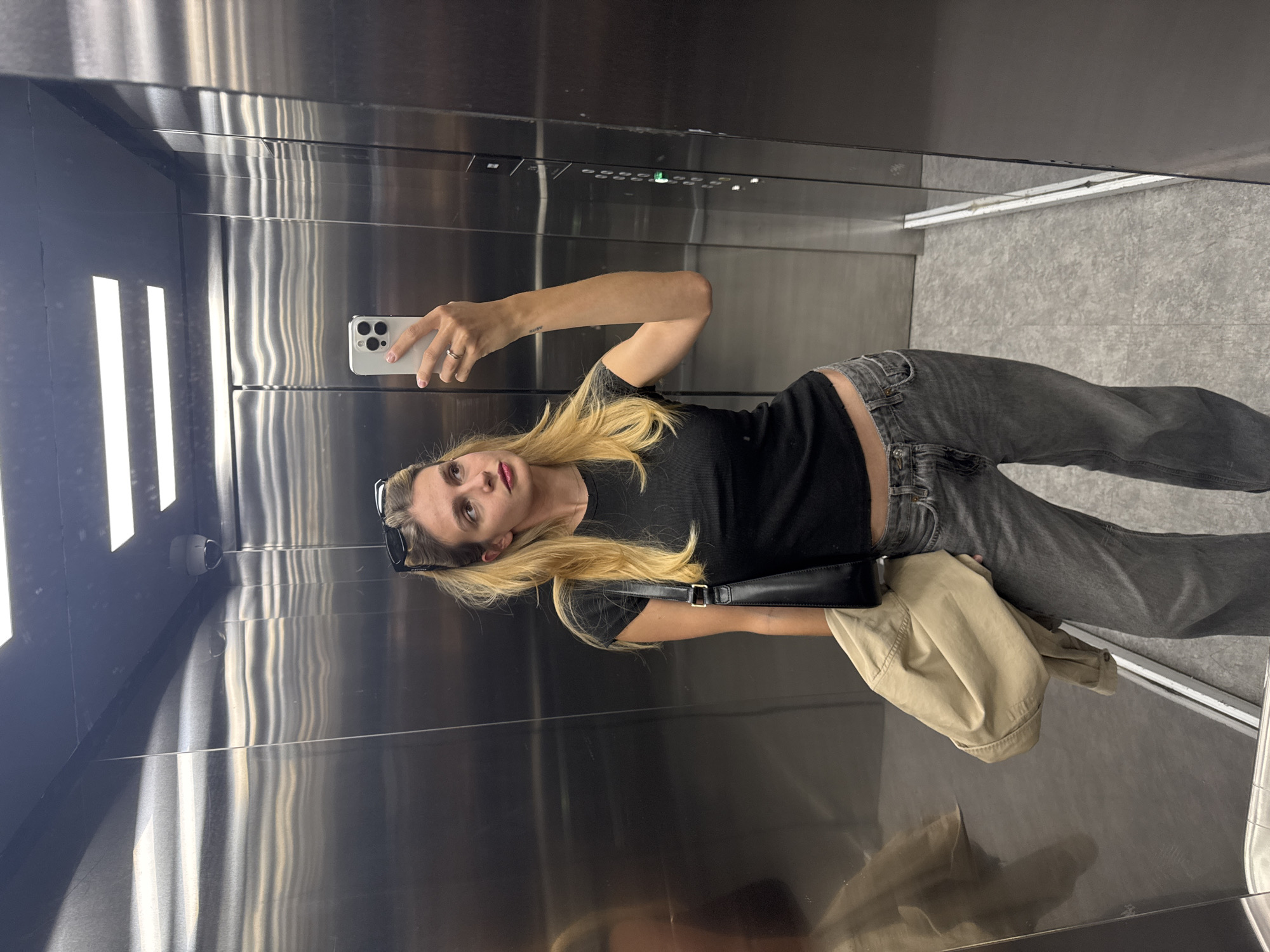 A woman is taking a selfie in an elevator. She is holding a smartphone in her right hand and looking at the screen. She has blonde hair, a black t-shirt, and grey jeans. A black bag is on her left side. A beige jacket lays on the floor next to her. The elevator has stainless steel walls, with a security camera and lighting fixtures visible. The floor is light grey. The location is unknown.
