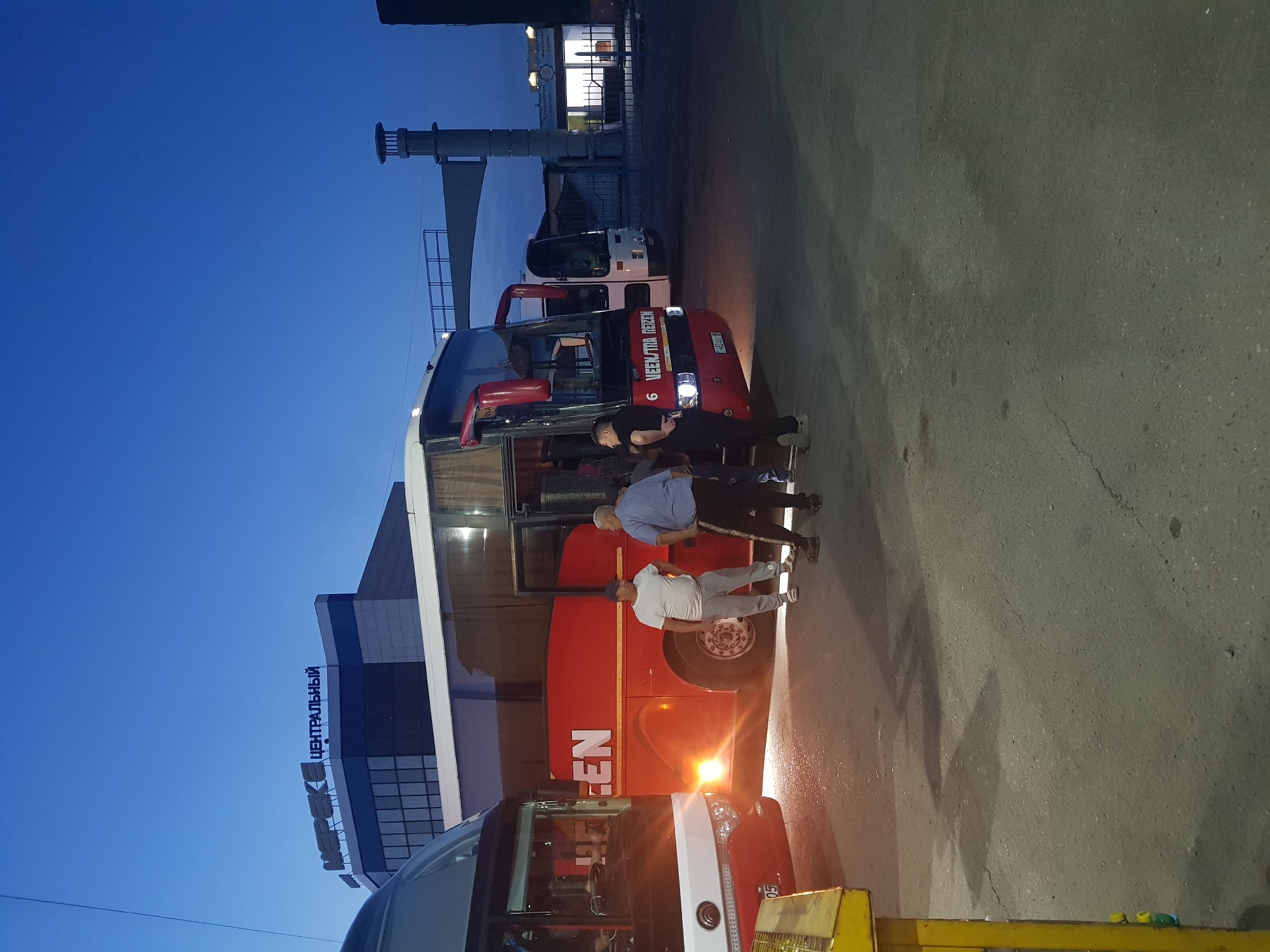 The image depicts a scene at what appears to be a bus station in the city of Semey, Kazakhstan, during twilight or early evening. Several buses are parked on an asphalt surface. In the foreground on the left, the front of a red and white bus is visible, with its headlights on. To its right, a large red bus is parked, featuring the white lettering "EEN" on its side. Three men are standing between the two buses. One man, wearing a white t-shirt and grey pants, stands facing the red bus. Another man, in a light blue plaid shirt and dark pants, stands talking to him. A third man, in a dark t-shirt and dark pants, stands with his back to the viewer, facing the red bus. In the background, a building with the Cyrillic text "ЦЕНТРАЛЬНЫЙ" (Central'nyy) and "МЕРЕКЕ" (Mereke) is visible. Further in the background, another bus and a dimly lit building with signage that appears to be in Cyrillic and possibly Kazakh script are present. The sky is a deep blue, indicating the low light conditions. The overall impression is of a bus departure or arrival point.