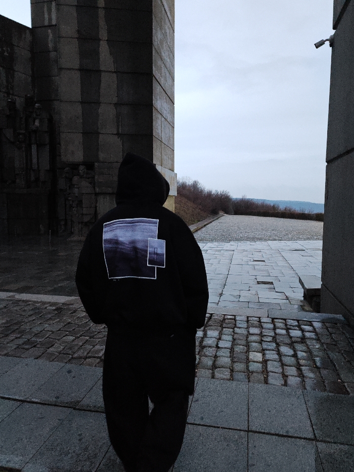 A single individual is observed from behind, positioned on an outdoor paved surface at the Monument to 1300 Years of Bulgaria in Shumen, Bulgaria. The person is wearing a black hooded sweatshirt featuring a graphic design on the back, consisting of two overlapping rectangular elements: a larger one displaying a blurry, dark, horizontal landscape or abstract pattern, and a smaller, slightly offset one containing a vertical dark line. Dark trousers are also visible.
The subject stands facing away from the camera, looking towards an open vista. To the left, a massive, grey concrete brutalist structure, part of the monument, exhibits vertical segments and sculpted relief figures. To the right, another simpler, grey concrete pillar or wall rises vertically, with a white security camera mounted near its top, facing into the scene.
The ground directly beneath and in front of the individual consists of light grey square paving stones, transitioning into smaller, dark grey cobblestones. Beyond the immediate foreground, a wide, open paved area extends into the distance, bordered by low, bare vegetation on a sloping embankment. The background reveals distant hills under an overcast, light grey sky. The overall lighting is diffused, indicating an overcast daytime condition.