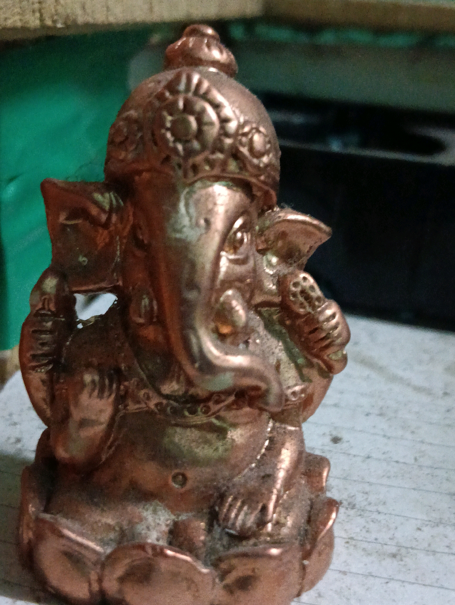 A close-up, eye-level, medium shot captures a small, metallic statue of Ganesha, the Hindu deity. The statue is bronze-colored and sits on a flat, textured surface that appears to be a paper with faint lines. The background is blurred, but shows a dark, horizontal object on the right and a green, textured surface on the left. The statue is intricately detailed, with a crown, an elephant-like face, and several arms holding objects. The lighting is dim, creating shadows and highlighting the metallic sheen of the statue. There is no discernible location within the city of South Dumdum, India, visible in the image; the scene appears to be an indoor setting.