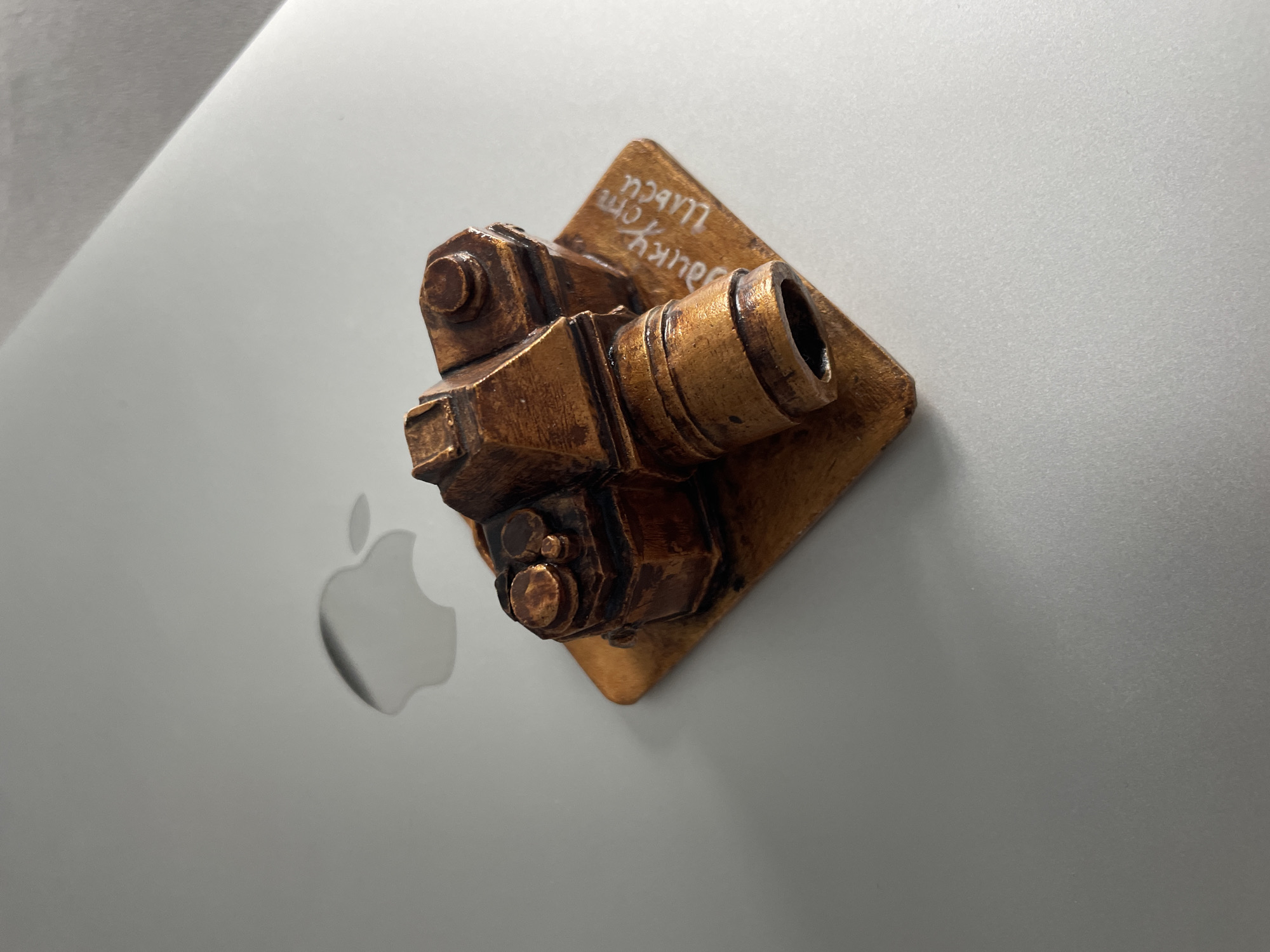 A miniature replica of a vintage camera, made of a bronze-colored material, rests on a square wooden base. The base is placed on a silver laptop, visible in the background with a subtle Apple logo. White lettering is inscribed on the wooden base, partially obscured by the camera. The scene is lit from above and to the right, casting a soft shadow to the left of the camera. The background is a smooth, light gray surface, likely the laptop lid. The image is taken from a high-angle perspective.