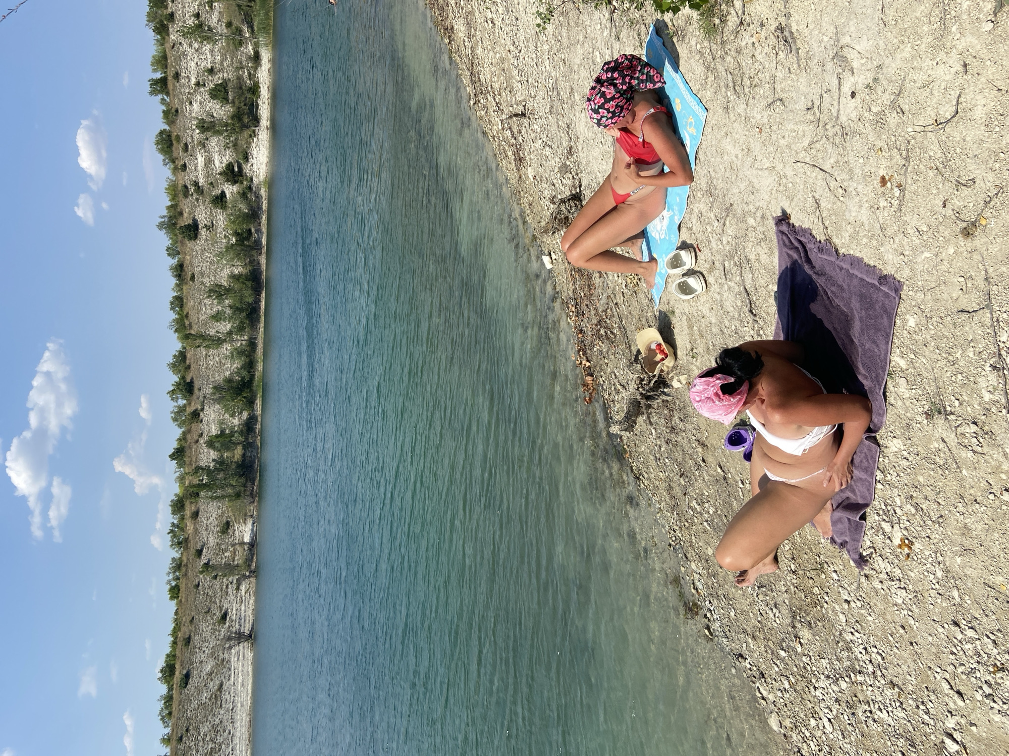 Two individuals are sunbathing on a pebbled shore next to a calm, turquoise lake. The person in the foreground, on the left, is lying on their side on a purple towel, wearing a white bikini and a pink headscarf. The person on the right is lying on their back on a blue towel, wearing a red bikini and a black and pink floral headscarf. To their left are a straw hat with cherry tomatoes, and a pair of white slip-on shoes. In the background, a treed embankment rises from the shore. The sky above is bright blue with scattered white clouds.