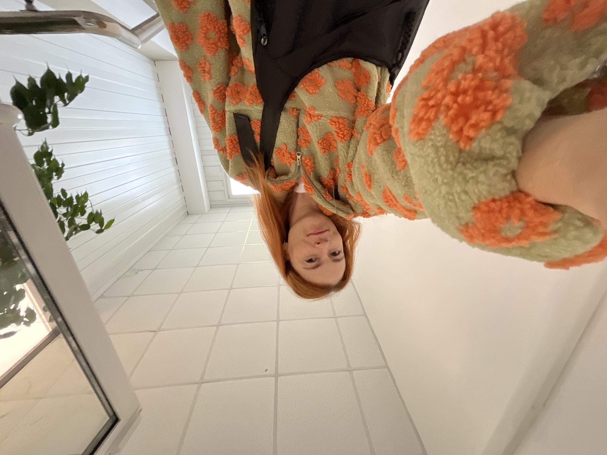 The media file is a selfie taken from a low angle, with the subject upside down. The subject is a person with long, reddish-brown hair, wearing a light-colored fuzzy jacket with an orange pattern. They have fair skin and are looking directly at the camera with a neutral expression. The background consists of white tiled walls and floor, suggesting an indoor setting such as a bathroom or a hallway. A plant with green leaves is visible on the left side of the image, near a window. The lighting appears to be artificial, and the time of day and weather are not discernible. There is no visible text in the image. The overall impression is a casual, personal photograph. The location context is Chaikovsky, Russia, but no specific landmarks or cues within the image confirm this.