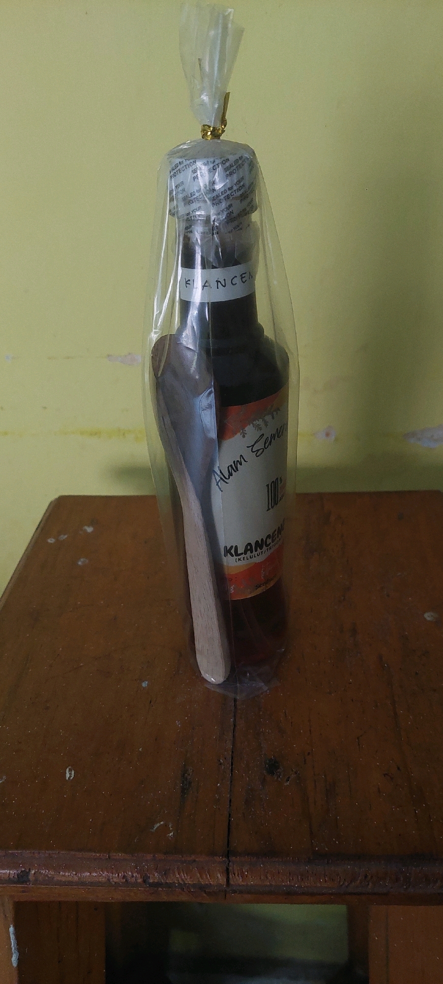 The image shows a bottle of "Alam Sema 100% KLANCENG" with a wooden spoon inside, all wrapped in clear plastic and tied with gold ribbon. The bottle appears to be some kind of traditional medicine or health supplement, given the "KLANCENG" label, which refers to stingless bees, often used in traditional Indonesian remedies. The bottle has a label that reads "Alam Sema" and "KLANCENG". The background is a plain yellow wall. The bottle is sitting on a wooden surface, likely a table. The image appears to be taken indoors, with the lighting suggesting it might be daytime. The overall scene is simple and focused on the product.
