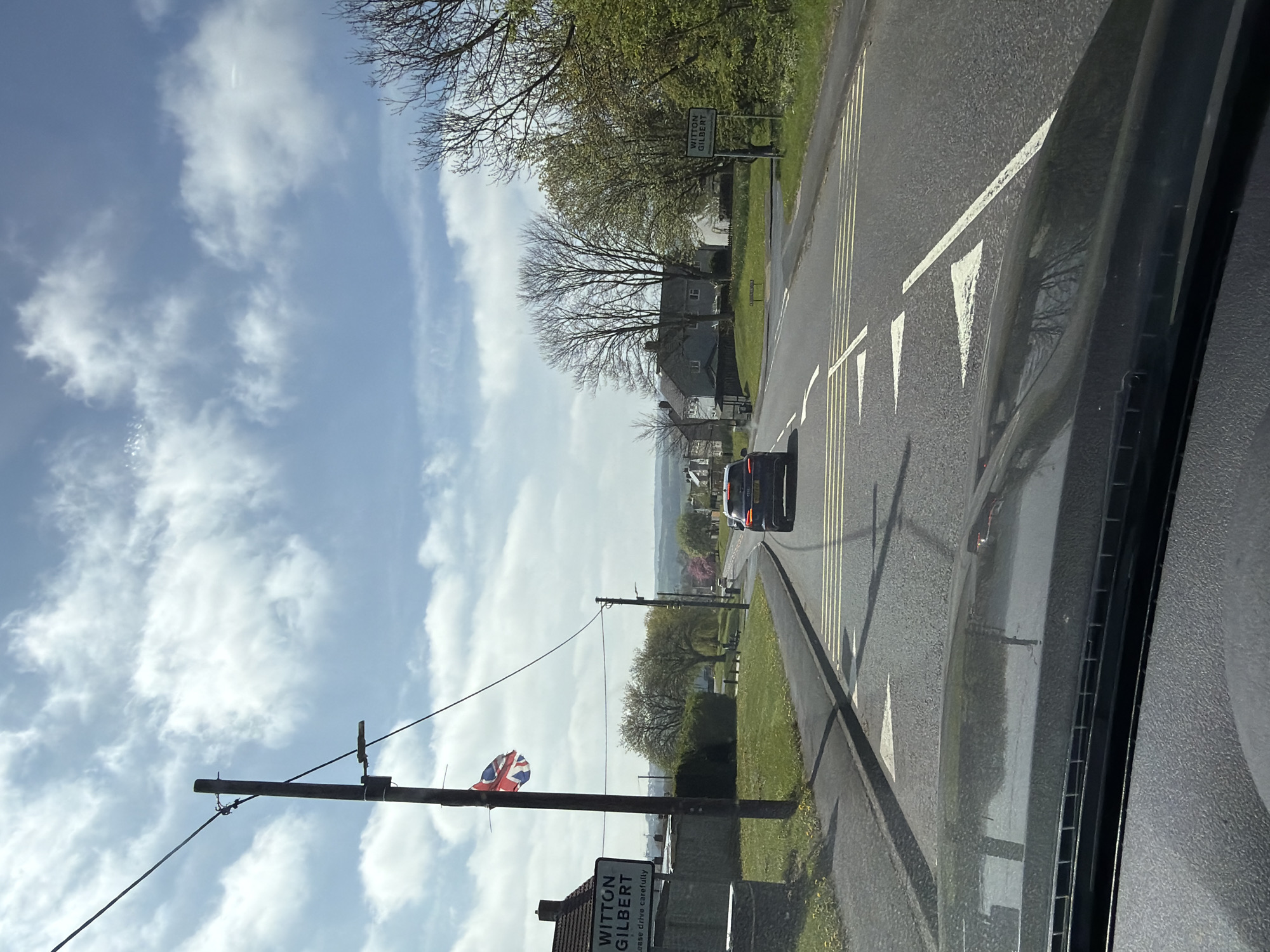 The image, taken from inside a vehicle, shows a street in Witton Gilbert, United Kingdom. The view is from the driver's seat, looking down the road. A car is visible ahead on the road. Road markings, including white triangles pointing inwards, are present. On the left side of the road, a sign for Witton Gilbert is affixed to a utility pole, which is also decorated with a Union Jack flag. The sky is visible with scattered clouds. Trees and vegetation line the roadside.