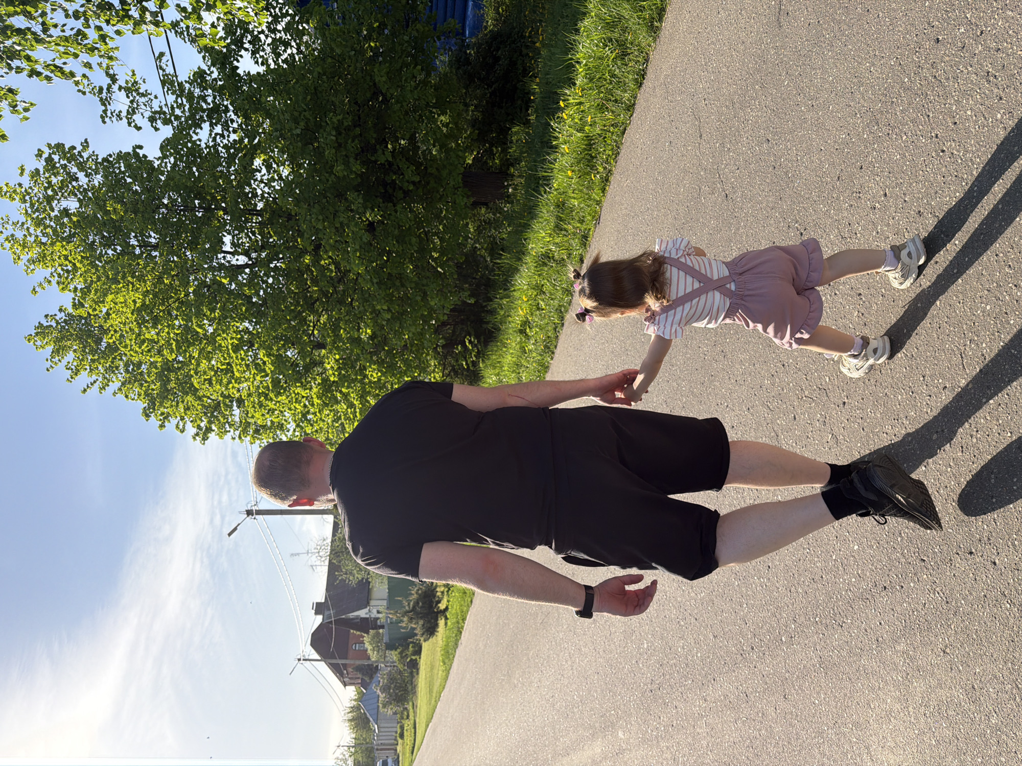 A man and a young child are walking hand-in-hand on an asphalt path. The man is wearing a black t-shirt, black shorts, black socks, and black sneakers. The child is wearing a striped top, pink overalls, white socks, and white sneakers. The path is adjacent to a grassy area and trees. The sky is visible and appears partly sunny. The location is Reshetnikovo, Russia, as indicated by previous context.