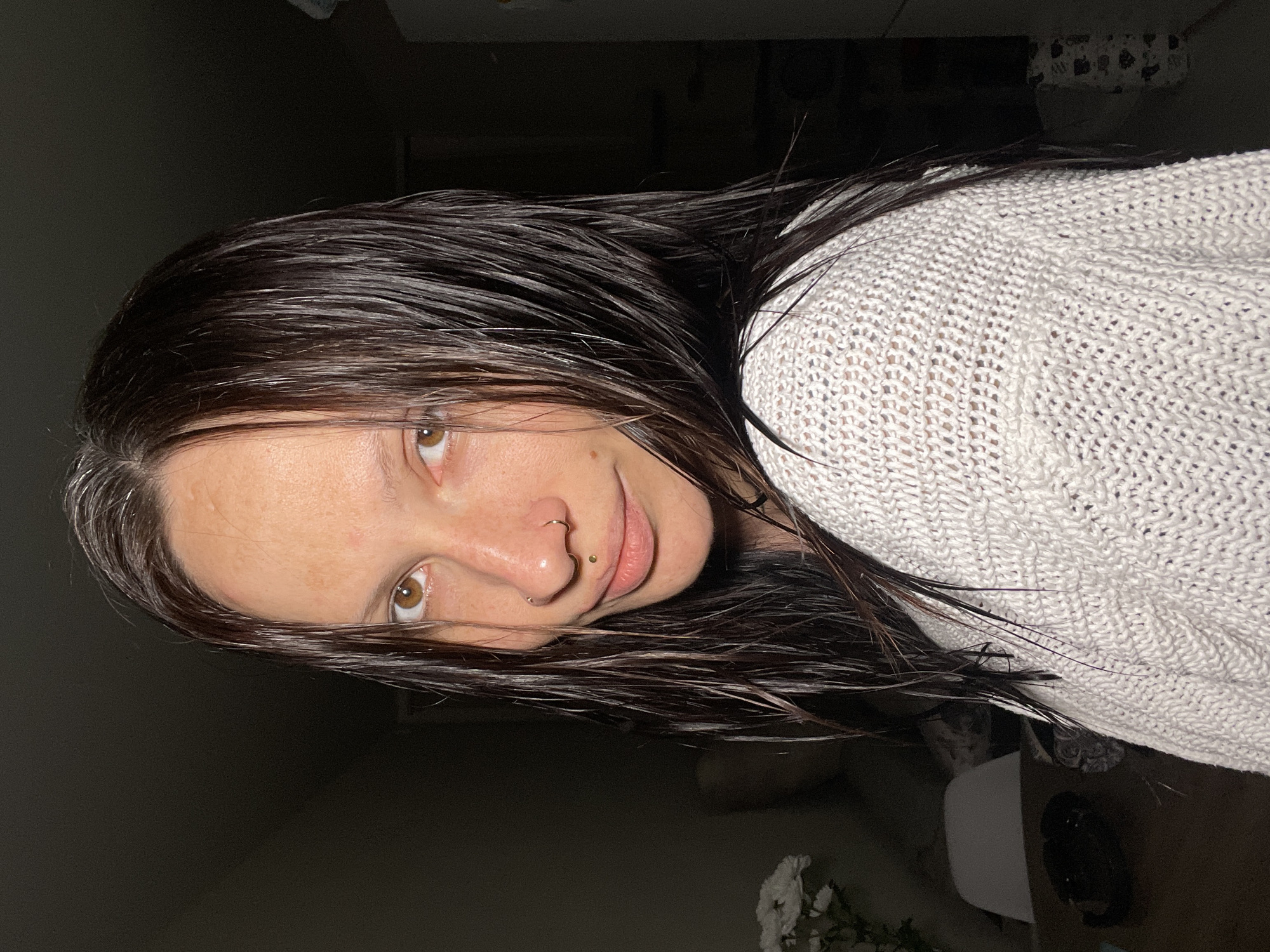 A woman with wet, dark brown hair and hazel eyes is facing the camera. She has fair skin with freckles and a nose piercing. She is wearing a white, knitted sweater. The background is dimly lit, revealing hints of a room interior with a vase of white flowers and furniture. The location of Pervomaysky, Russia is not discernible from the image.