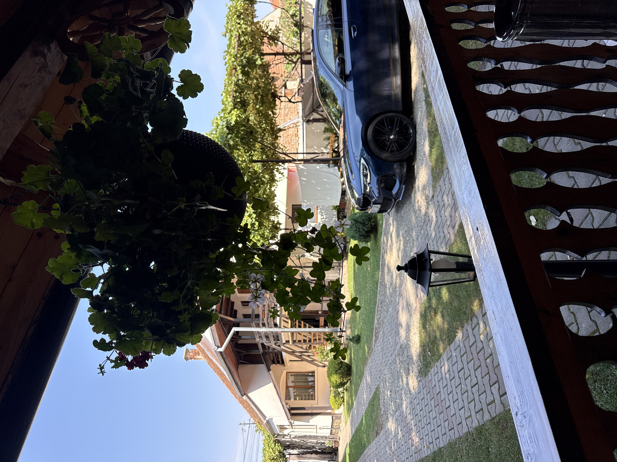 The image depicts a residential property in Cârțișoara, Romania. In the foreground, a dark wooden porch railing with decorative cutouts is visible. Beyond the railing, a blue BMW sedan is parked on a driveway made of interlocking pavers. The driveway is bordered by a well-maintained lawn and a pathway leading towards a two-story house. The house has a light-colored facade, a red tile roof, and wooden balconies. Green foliage, including a hanging basket of plants in the upper left, frames the scene. A black lantern stands beside the driveway. The sky is clear and blue, indicating fair weather. The overall scene suggests a peaceful, rural setting.
