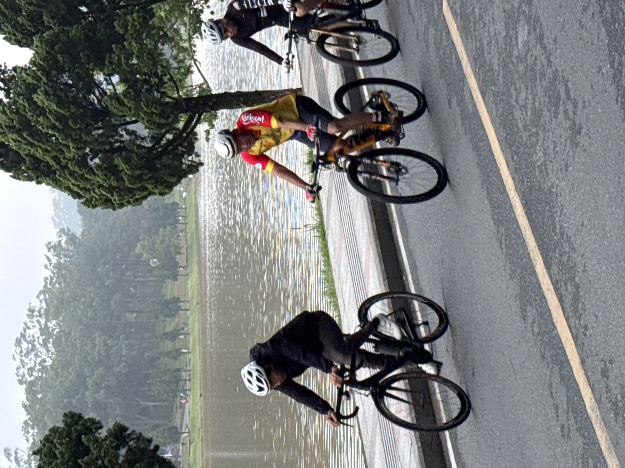 In Da Lat, Vietnam, three individuals are cycling on a paved road adjacent to a lake. The person in the foreground is wearing a black jacket, black pants, and a white helmet. They are riding a black bicycle with a water bottle attached. In the middle, a person in a yellow and red cycling jersey with black shorts and a white helmet is riding an orange and black bicycle. To the right, a person wearing a black cycling jersey with white accents and a white helmet is riding a black bicycle. The road has a yellow line marking its center. The background features a calm body of water, a sidewalk with some greenery, and a dense forest with tall trees. The weather appears overcast and possibly misty.